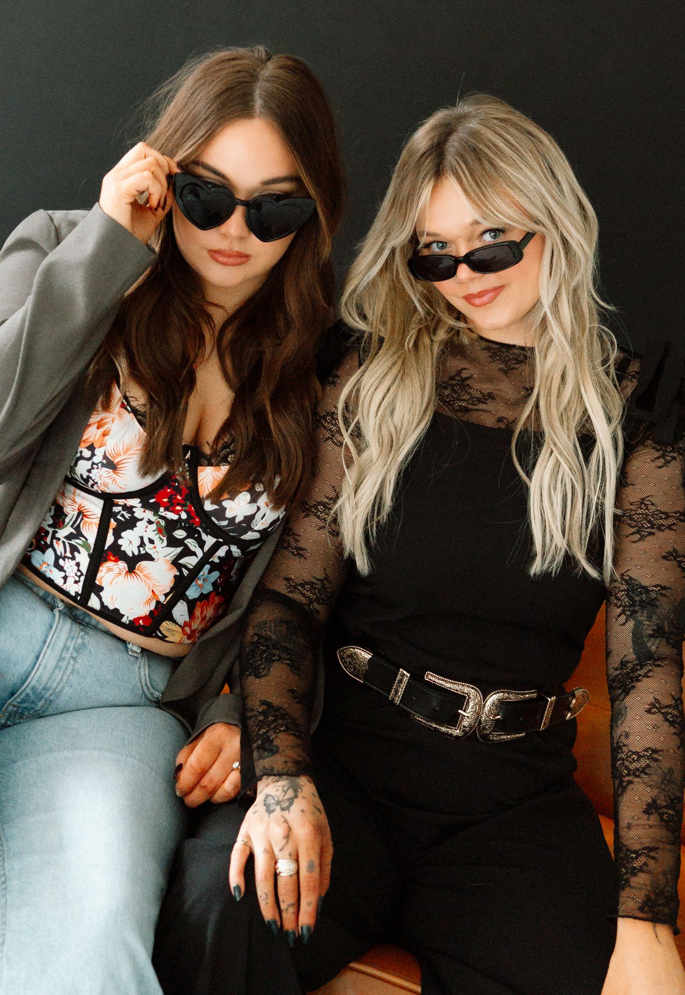 Two stylish women wearing black sunglasses pose together, one in floral top with jeans, other in black lace dress with belt.