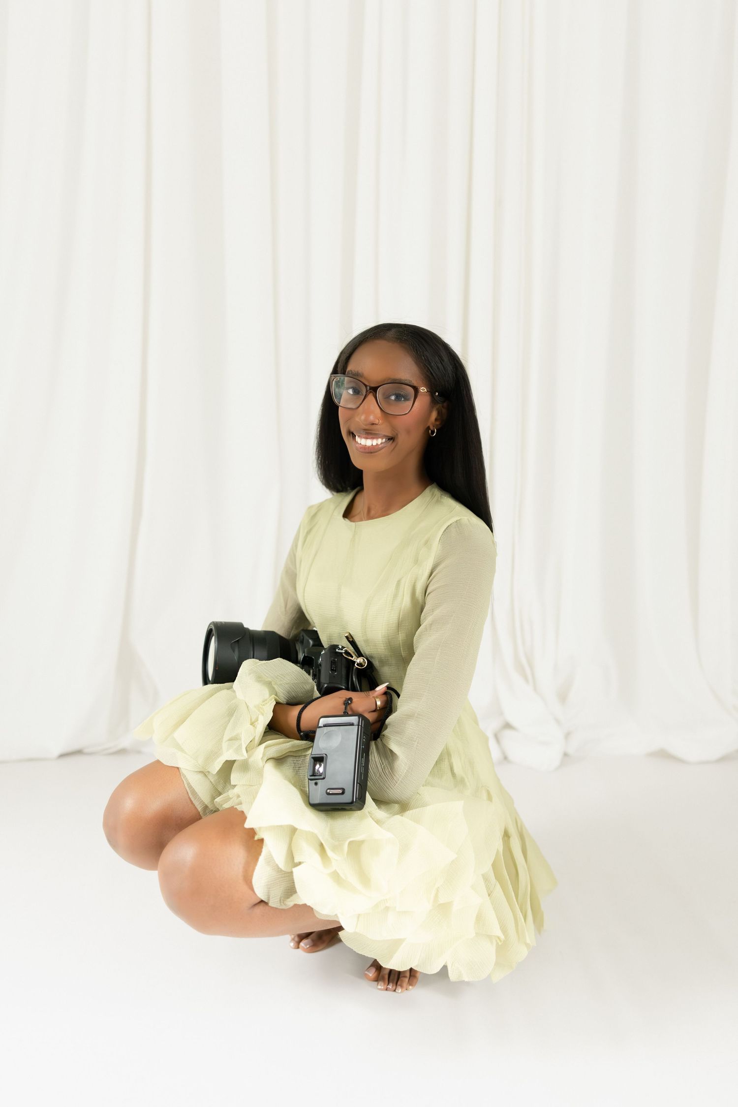 Young Black woman in green dress sitting on floor holding camera, smiling, white backdrop, glasses, studio portrait.