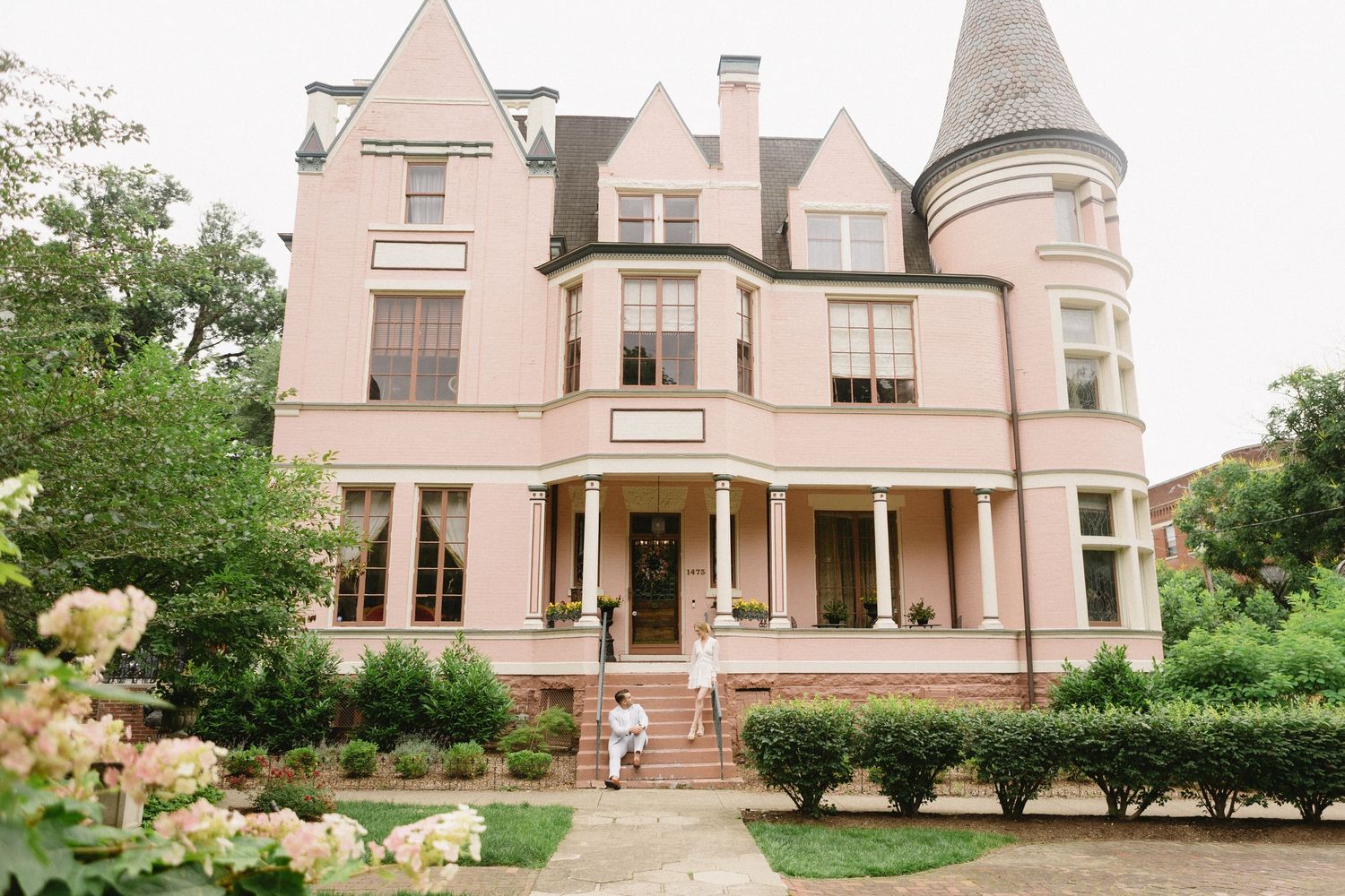 Elegant pink Victorian mansion with turret, lush gardens, and couple on steps.