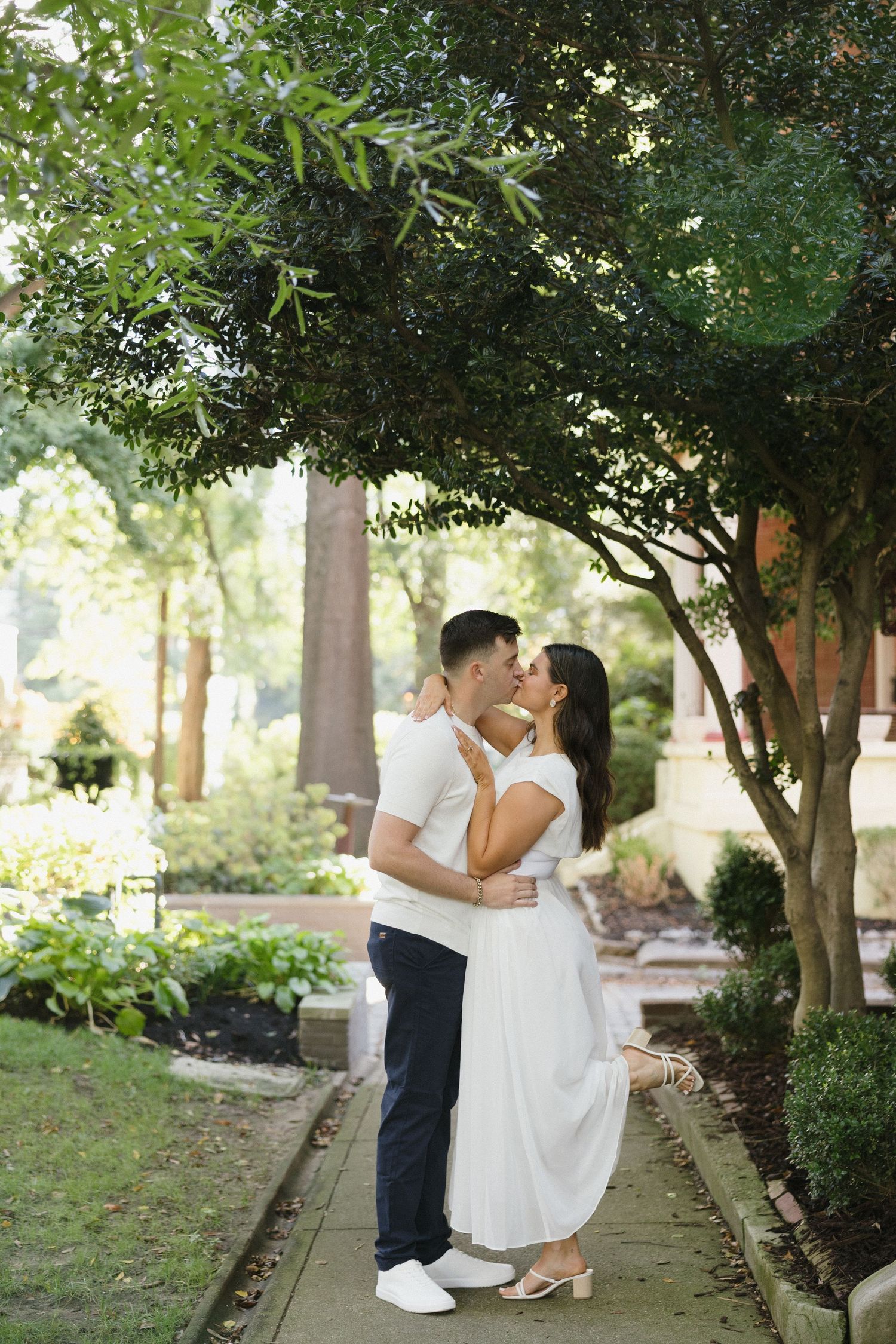 Couple sharing a kiss on a garden pathway surrounded by lush green trees and manicured shrubs.