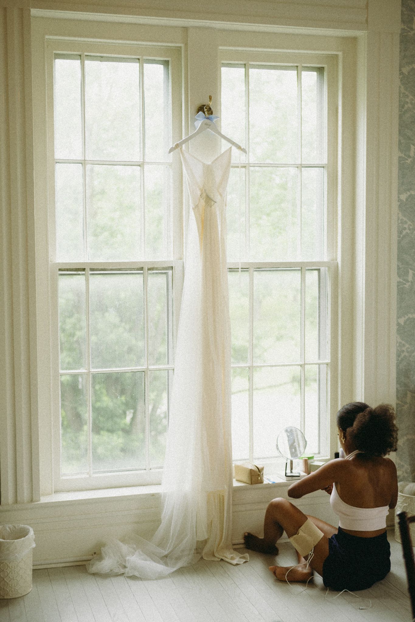 A white wedding dress hangs elegantly in front of tall windows while someone sits on the floor looking out at nature.