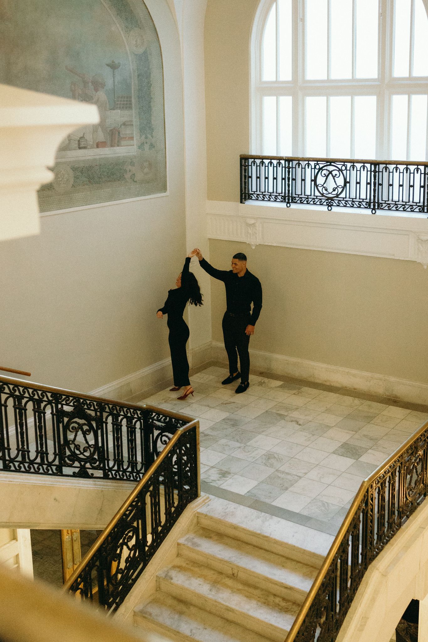 Two people dance together in an elegant stairwell with ornate black railings and marble floors.