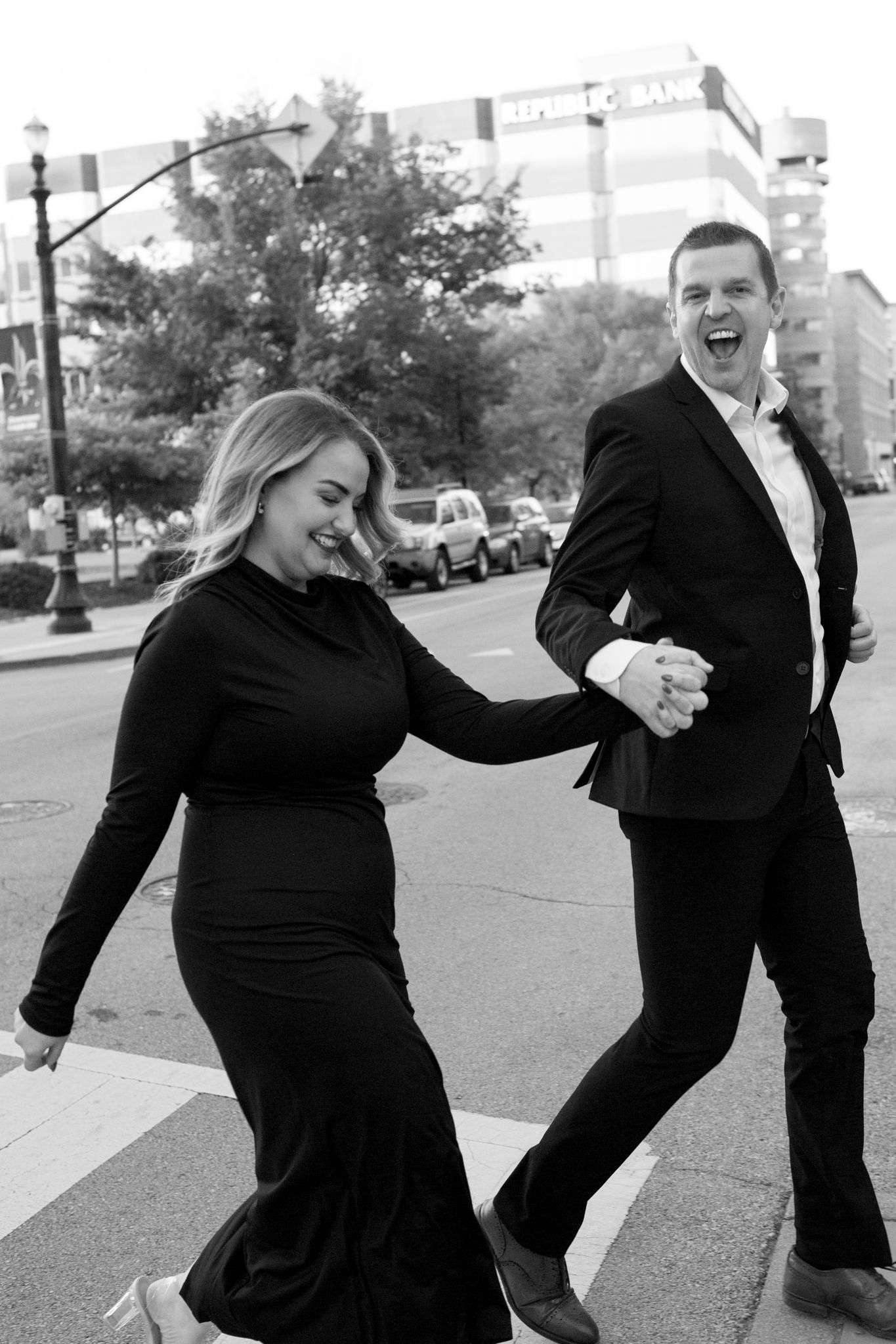 A couple in formal black attire share a joyful moment together while dancing on a city street.