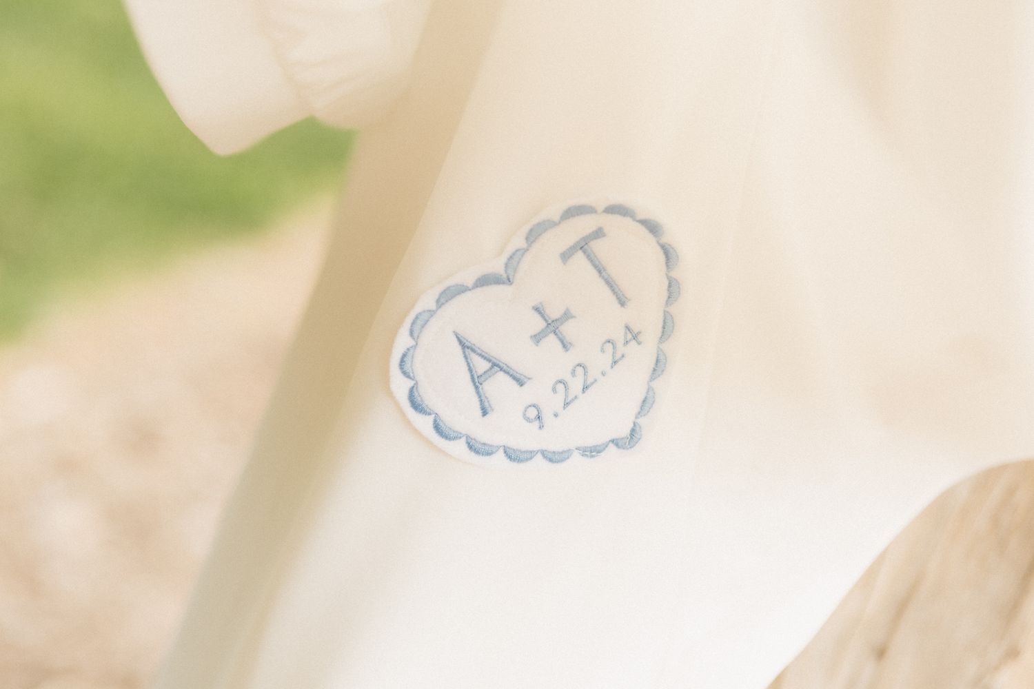 A delicate heart-shaped logo with initials embroidered in light blue thread on white fabric.