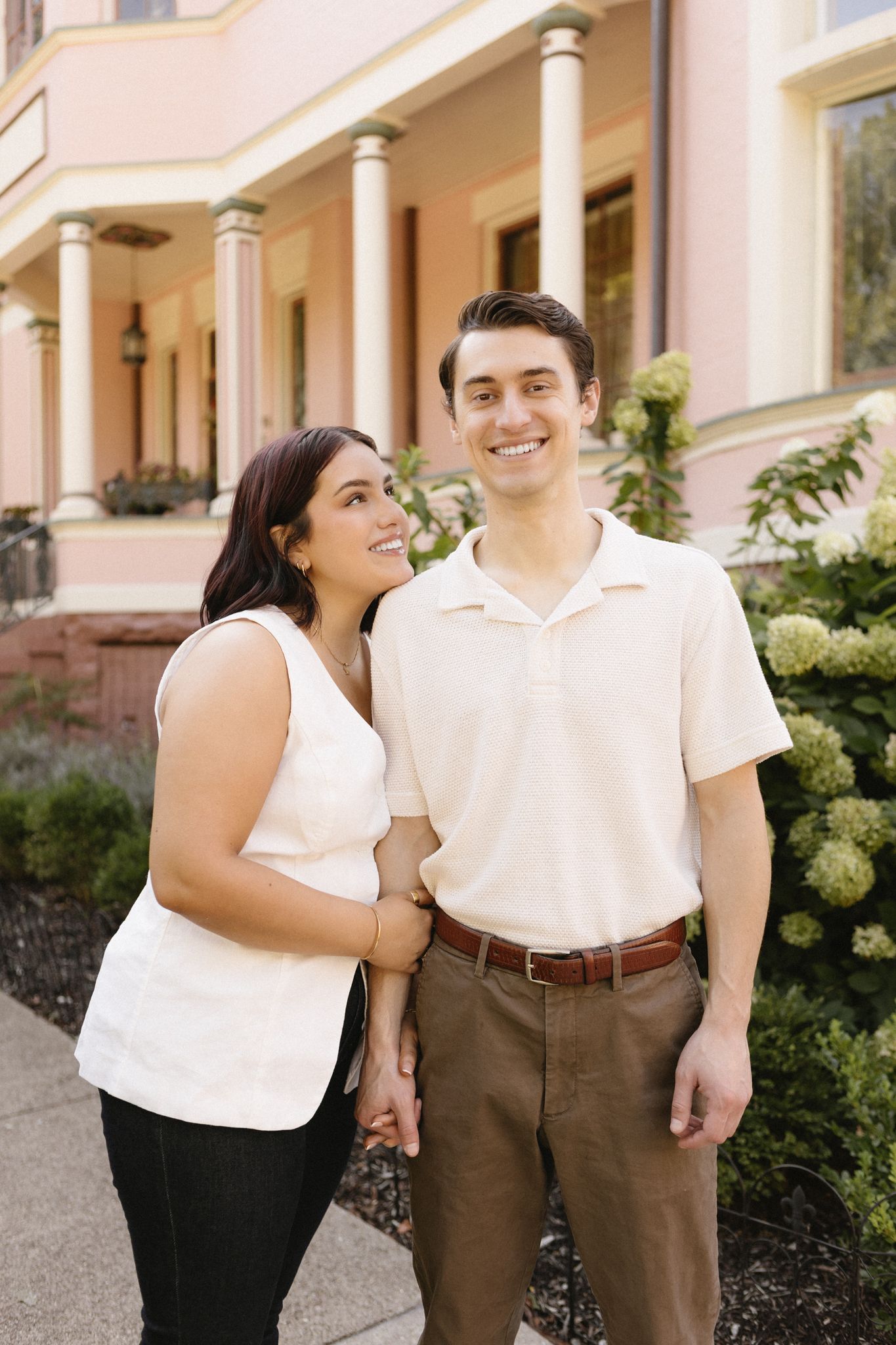 A couple in white and brown outfits smiling and embracing while standing outside a building with columns.