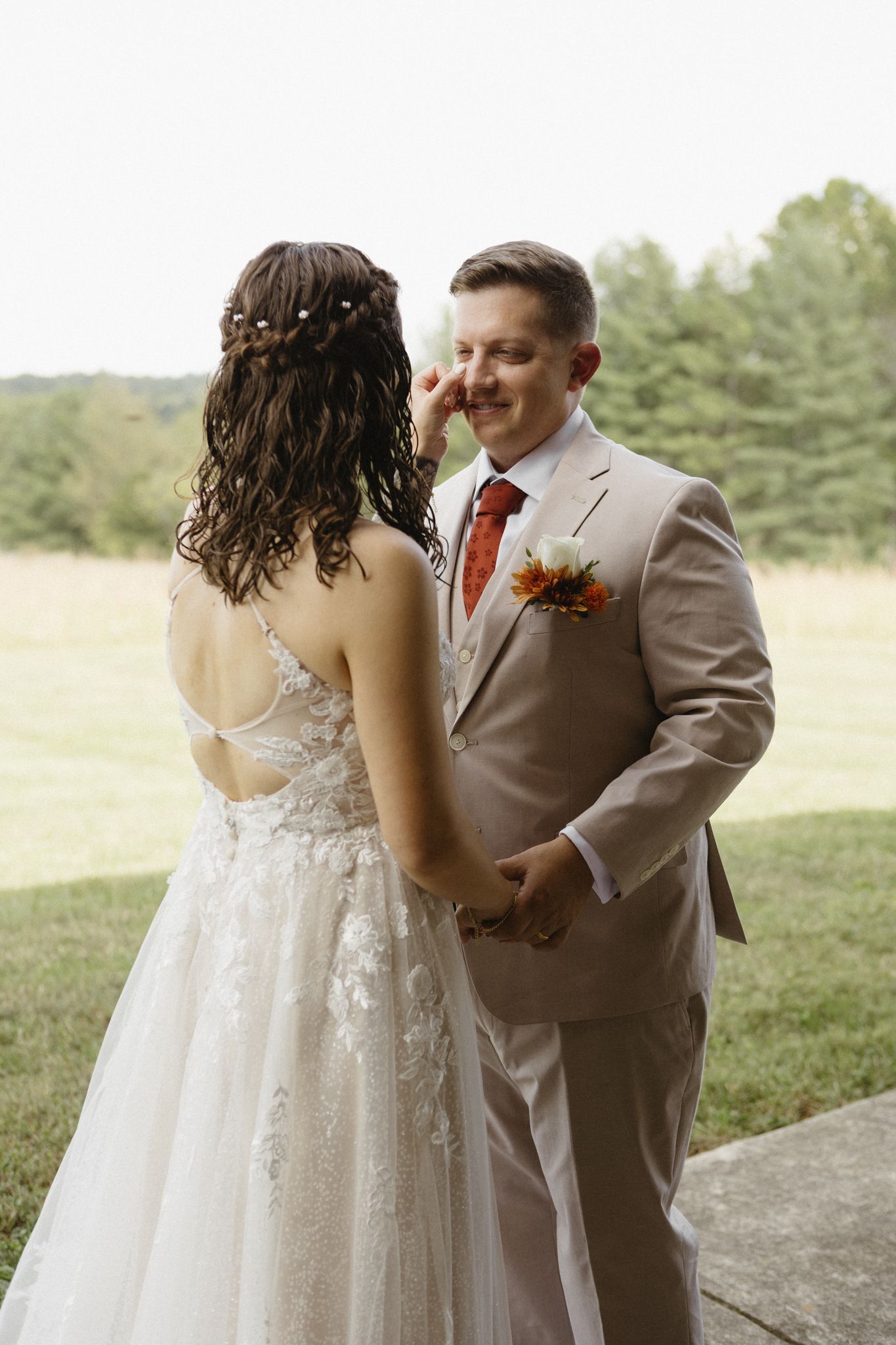 A couple shares an intimate first look moment outdoors with the bride wearing a lace gown and groom in beige suit.