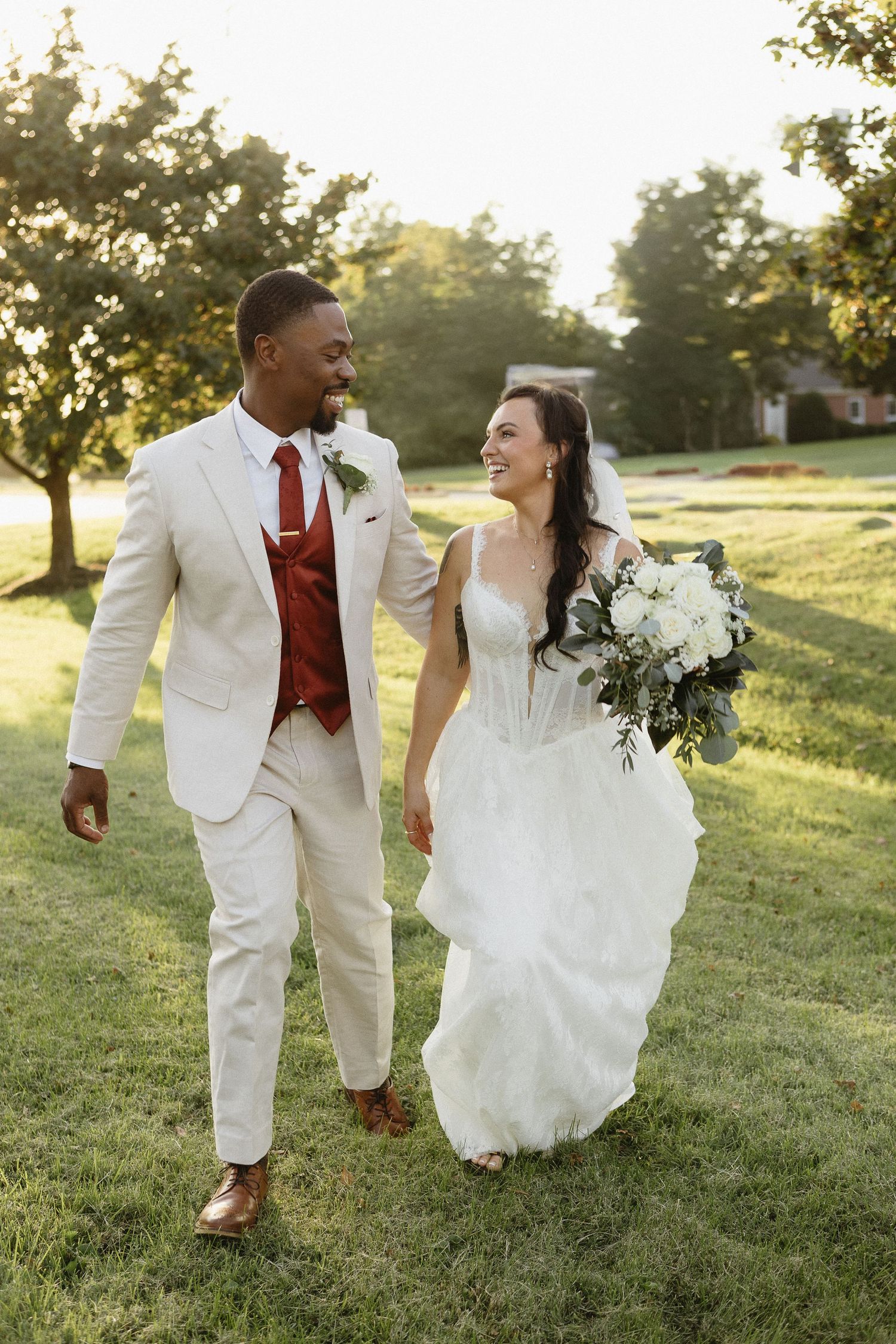 A joyful wedding couple walking together on grass during golden hour, with the bride holding a white bouquet.