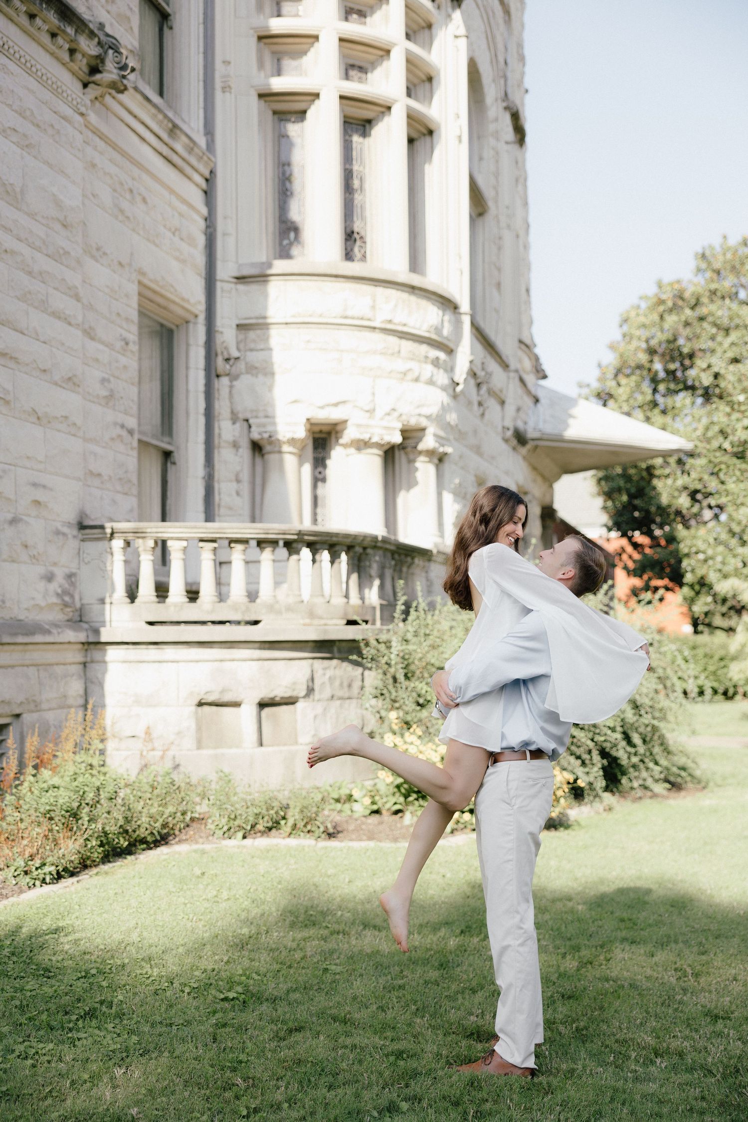 A joyful couple embraces outdoors, man lifting woman near an elegant stone mansion with lush gardens.