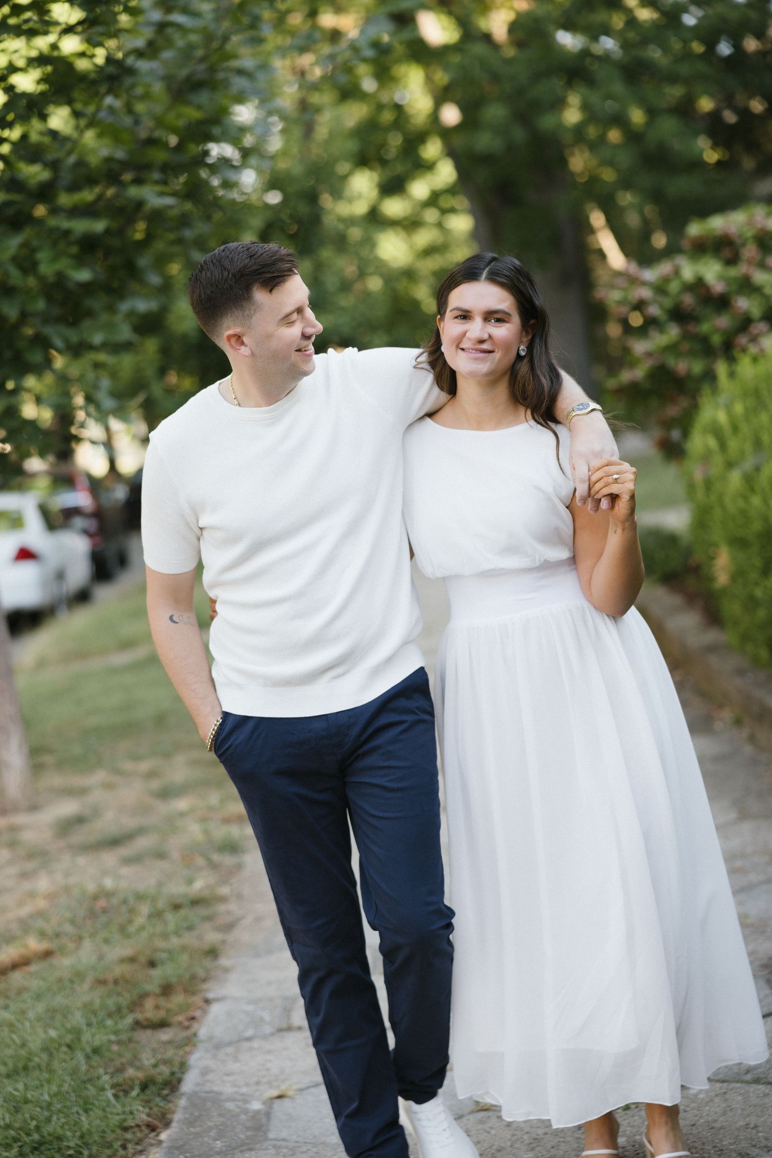 Happy couple walking together outdoors, man in white tee and navy pants, woman in elegant white midi dress, park setting.