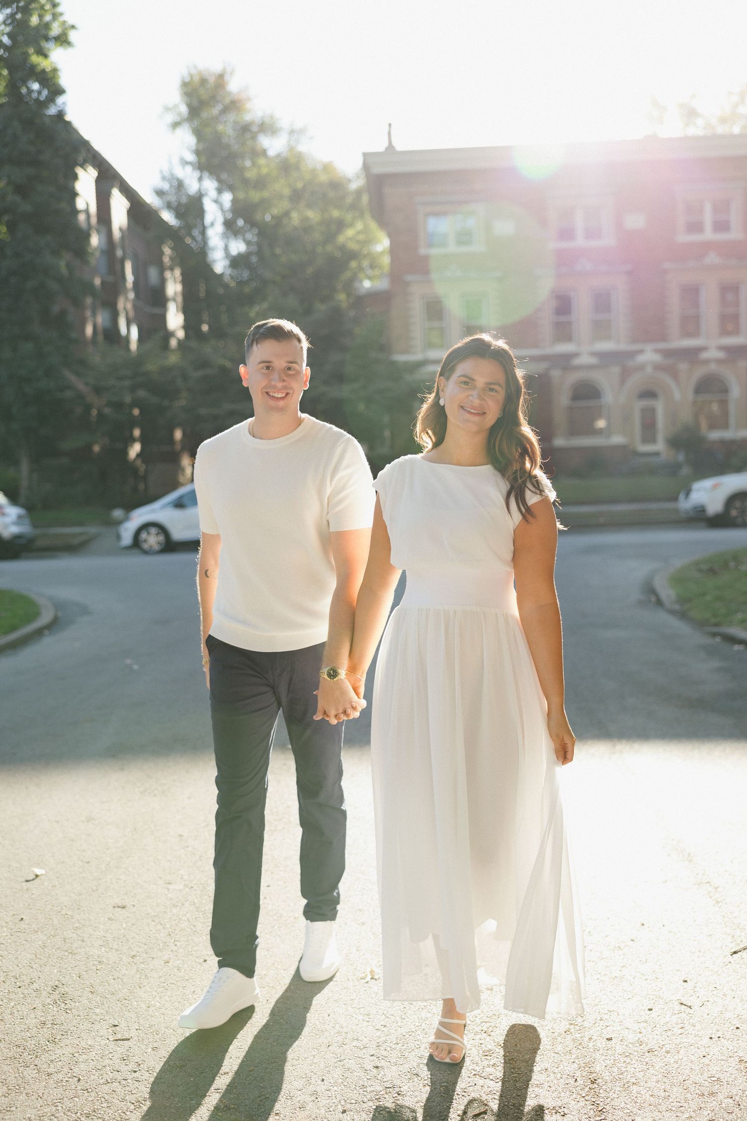 A smiling couple holds hands outdoors in golden sunlight, woman in white dress, man in white tee and gray pants.
