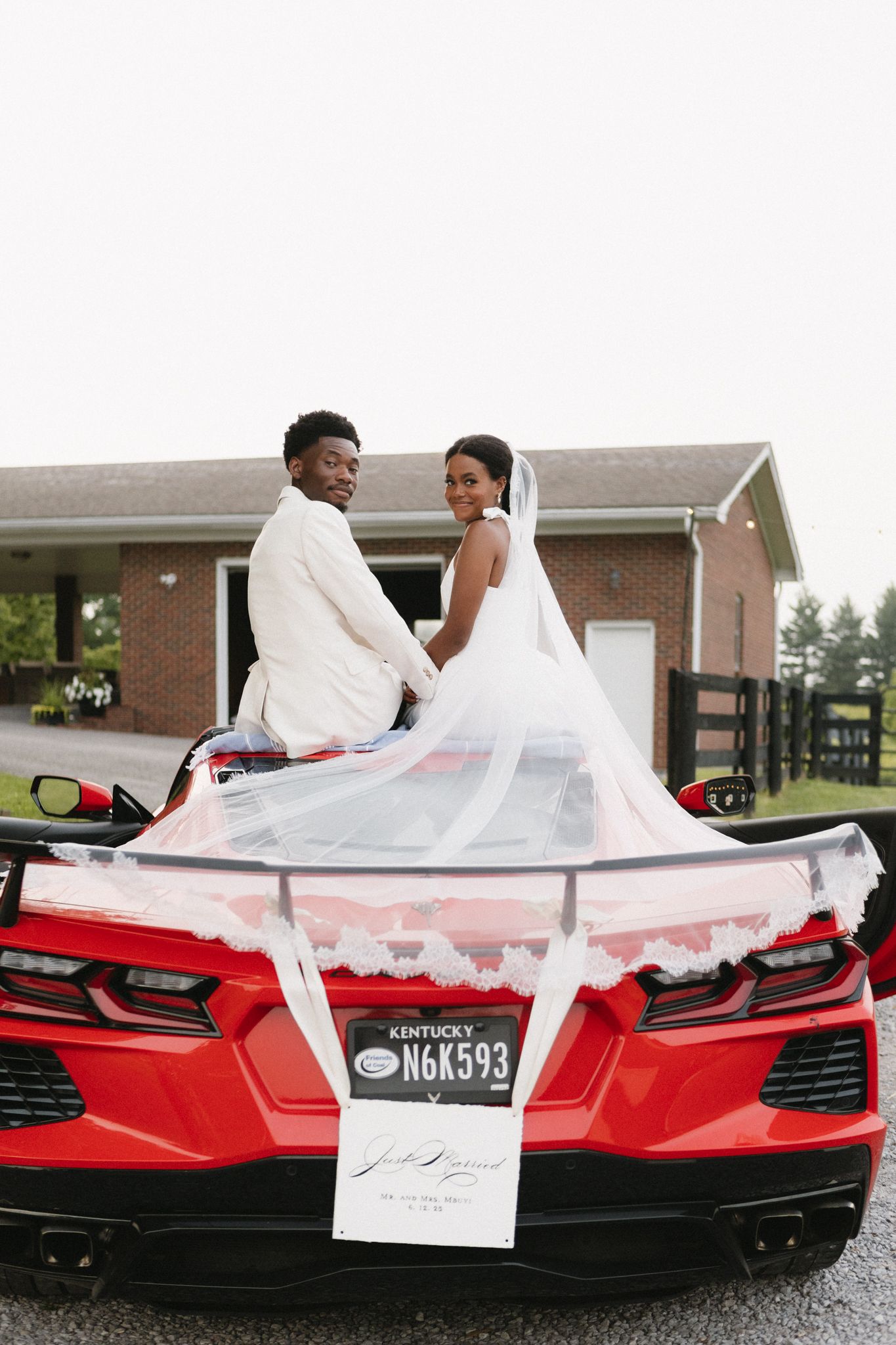Newlyweds pose on a striking red sports car decorated with wedding accents outside a brick building.