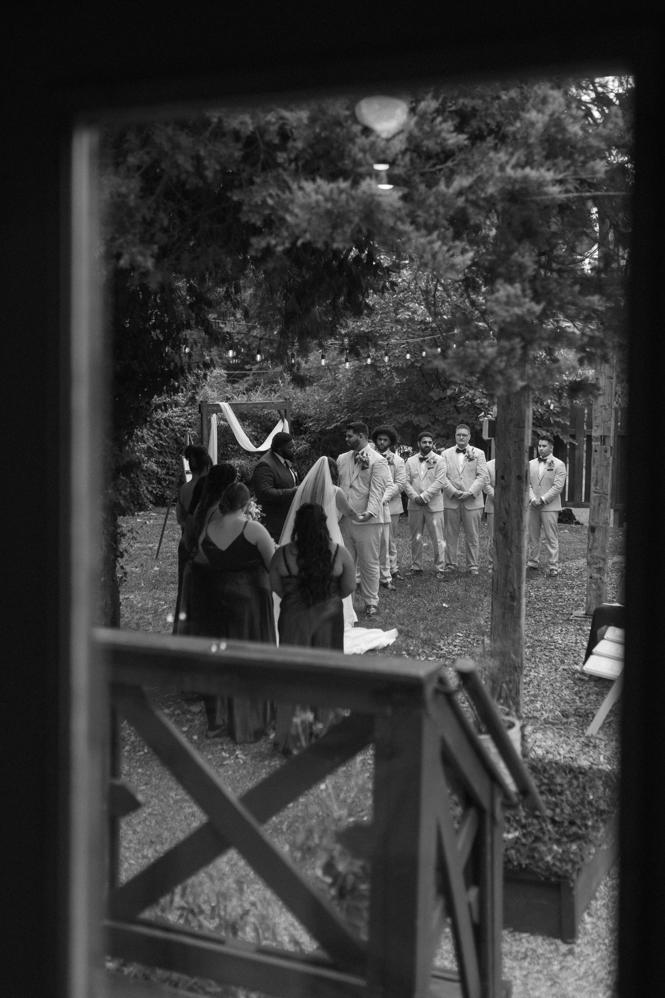 Black and white wedding party scene with harpists playing music as groomsmen in light colored suits approach on a garden path.