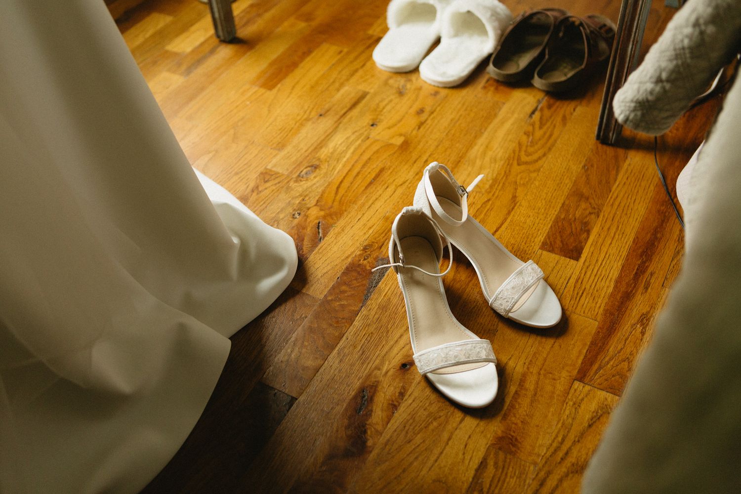 White strappy wedding sandals rest on a hardwood floor next to a dress hem and mirror reflection.