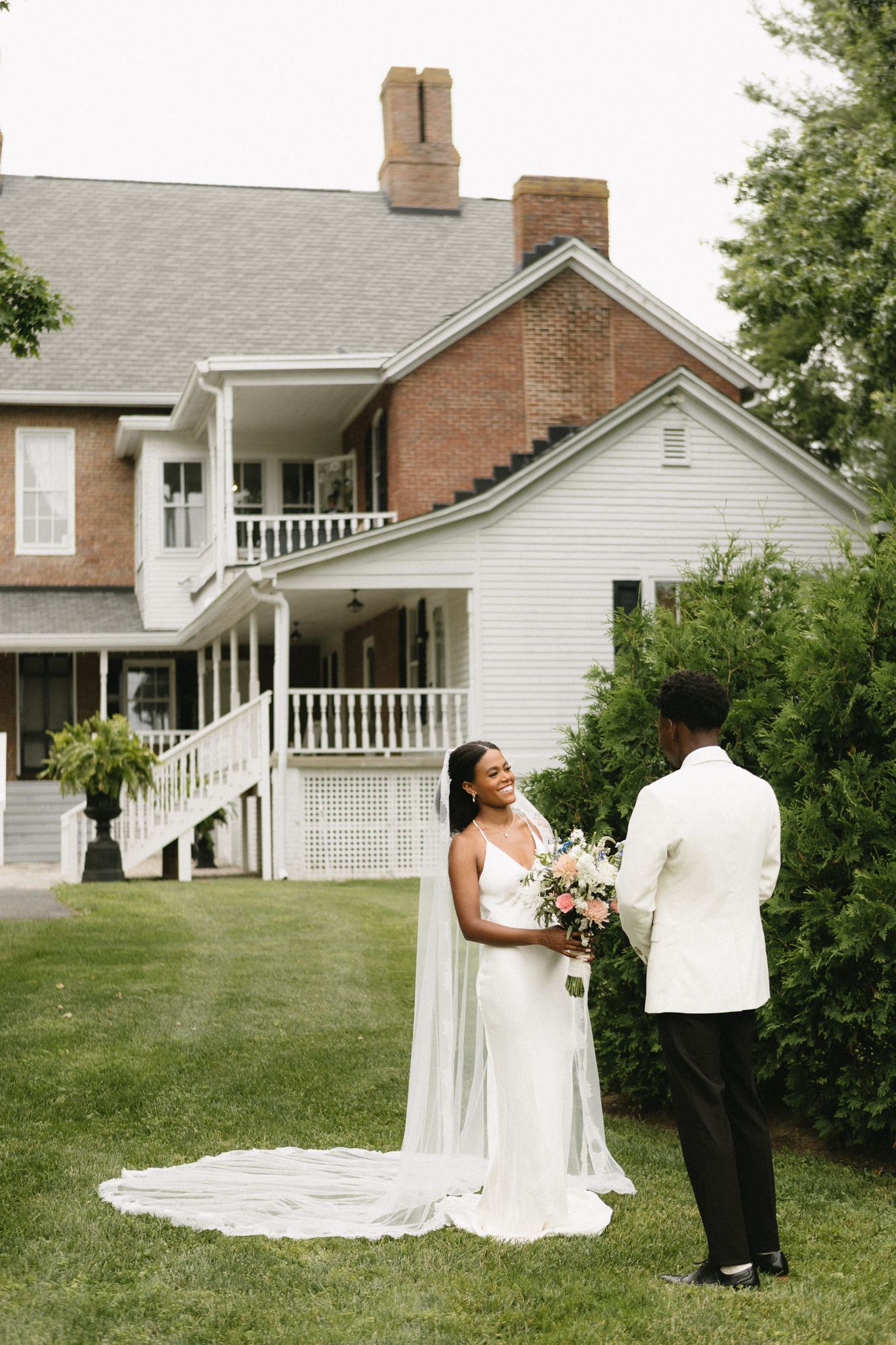 A wedding couple stands on a lawn in front of a beautiful brick and white farmhouse with wrap-around porches.