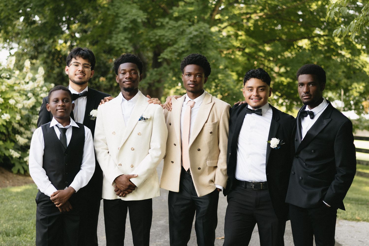 Groomsmen in formal tuxedos and tan suits pose together outdoors for wedding photos.