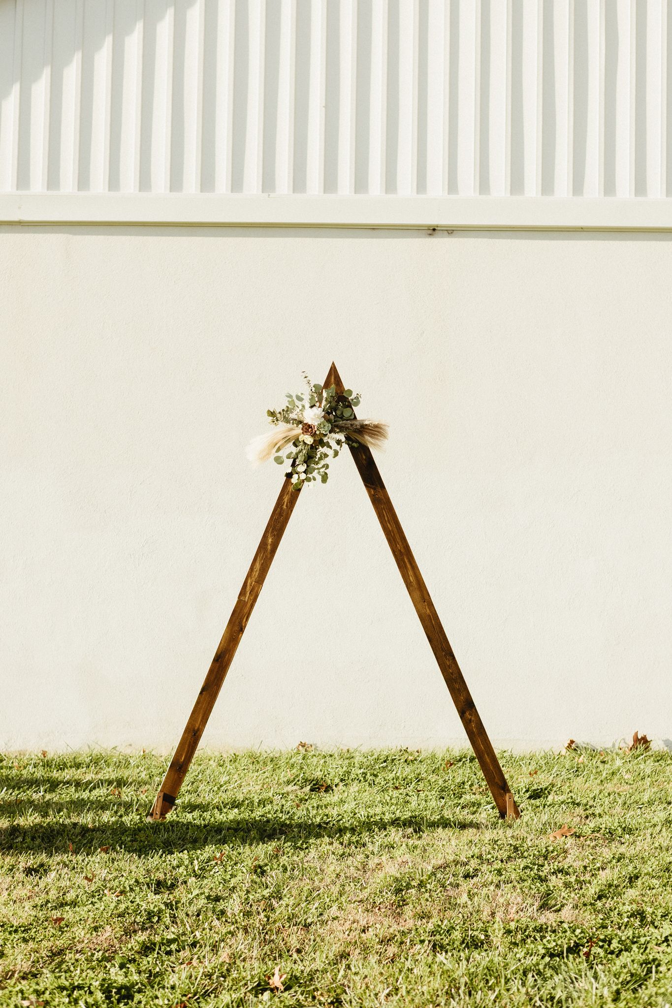 A rustic wooden A-frame structure with floral decorations stands on a grassy hillside during an outdoor event.