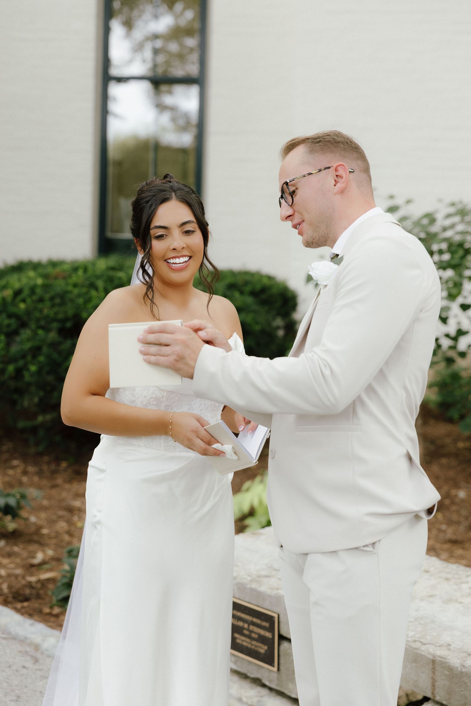 Smiling bride and groom in white attire exchanging handwritten vows outside on their wedding day.