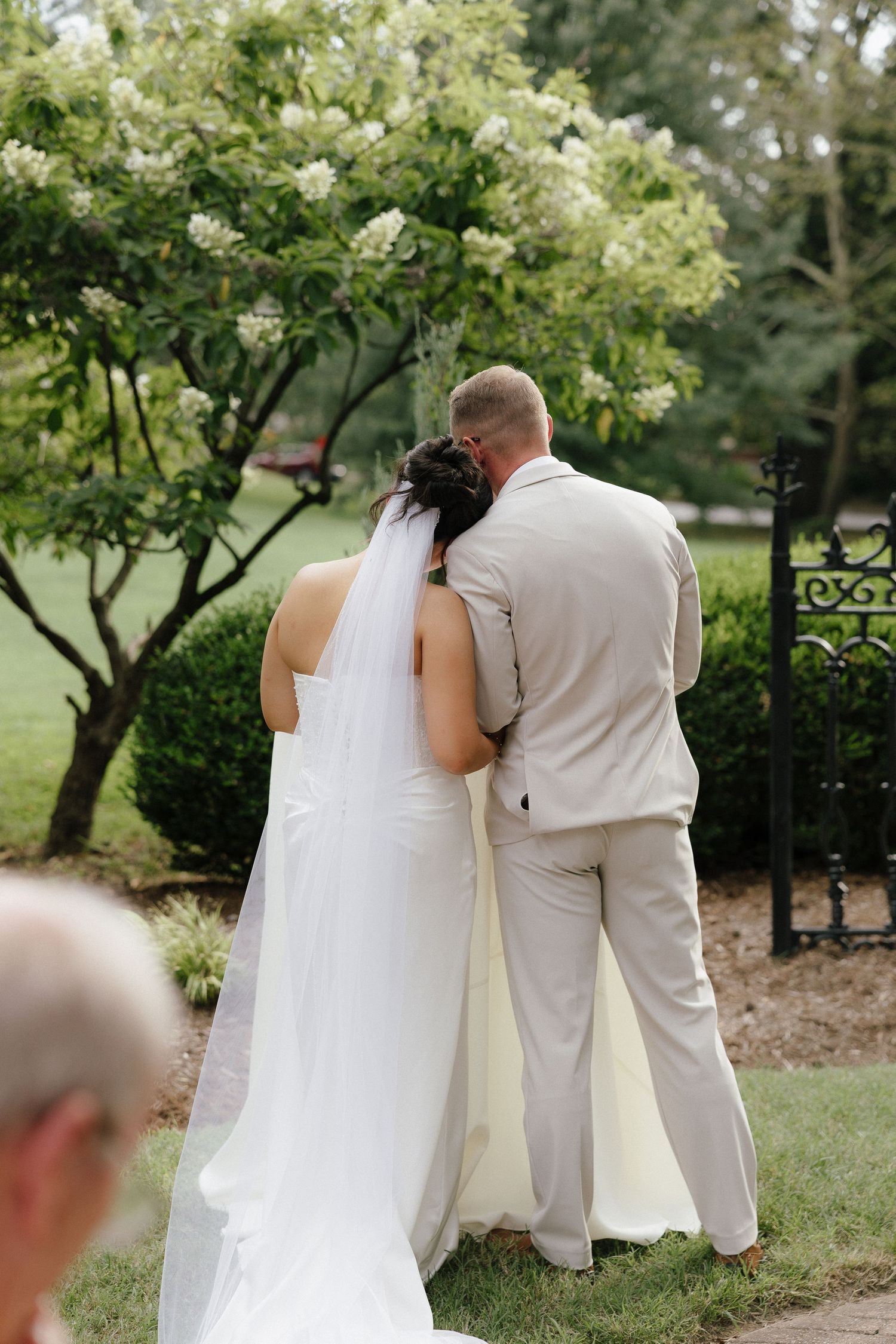 Bride and groom stand together from behind near blooming white tree in lush garden at outdoor wedding.
