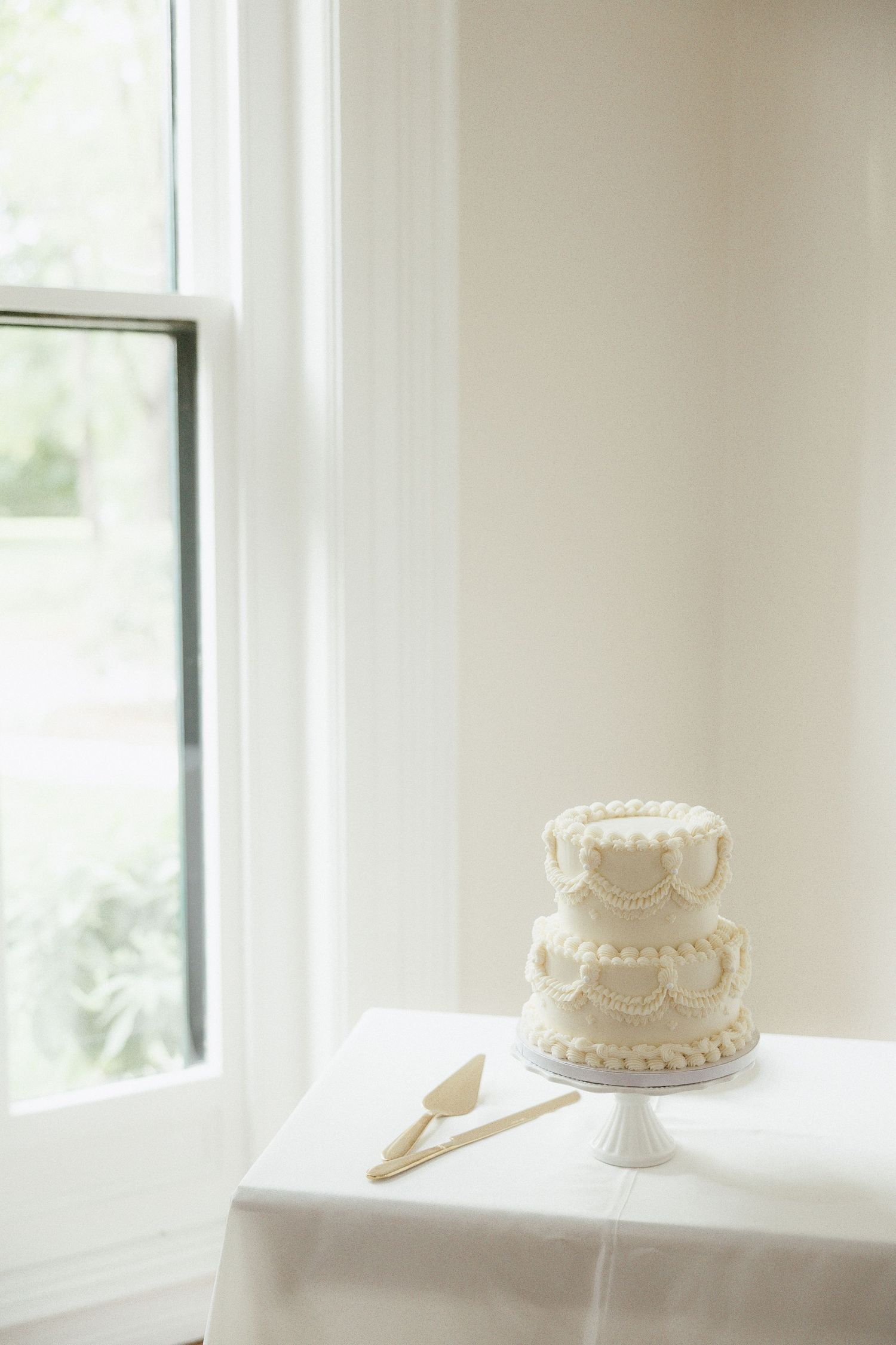 Two-tier white wedding cake with piped scallop details on a white pedestal stand beside a window.