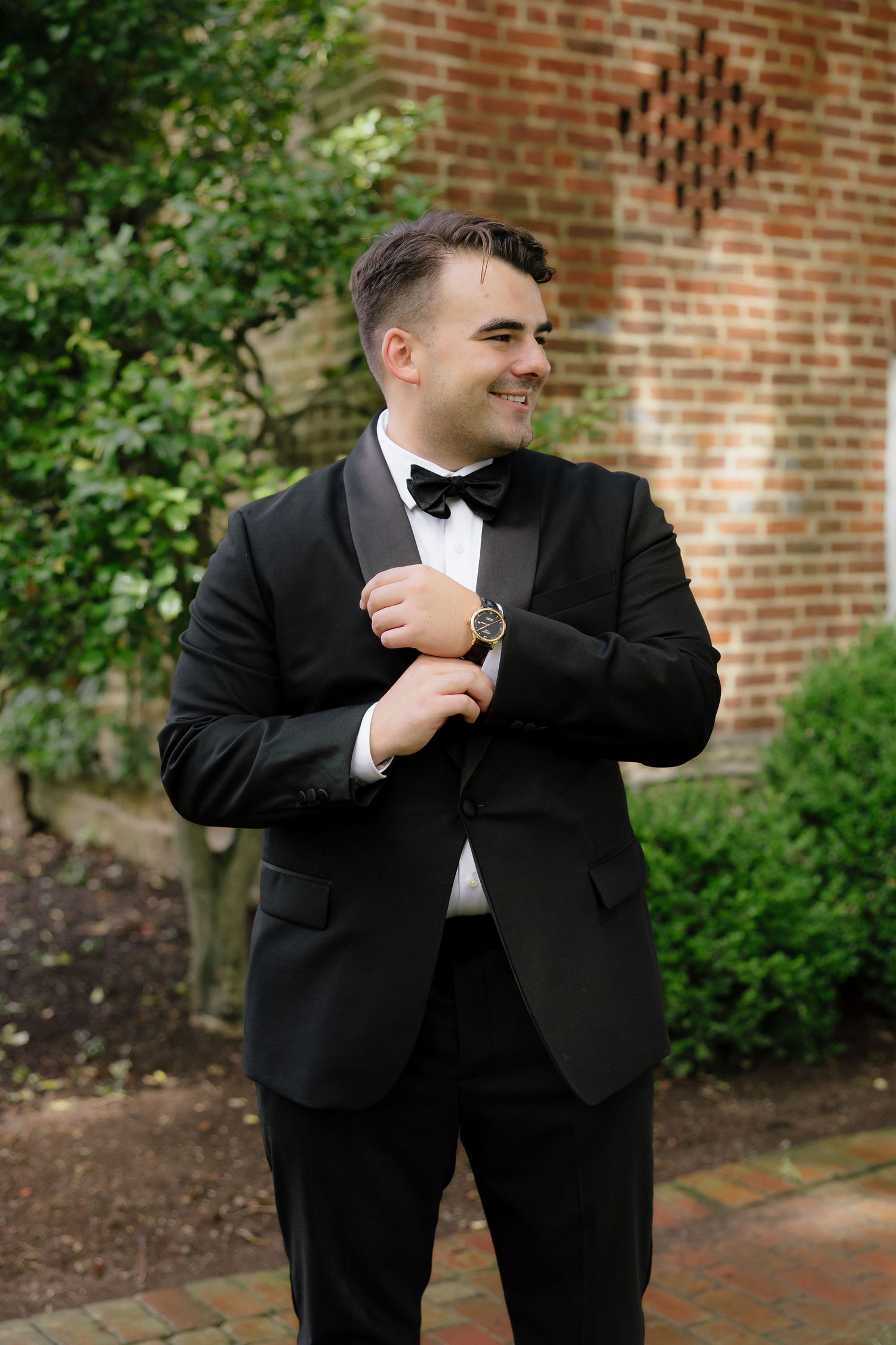 Dapper groom in black tuxedo and bow tie adjusting cufflinks outdoors near brick wall and green shrubs.