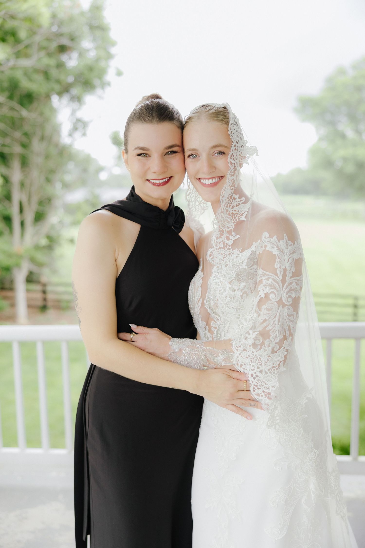 Bride in lace wedding gown and veil poses with bridesmaid in black dress on outdoor porch.