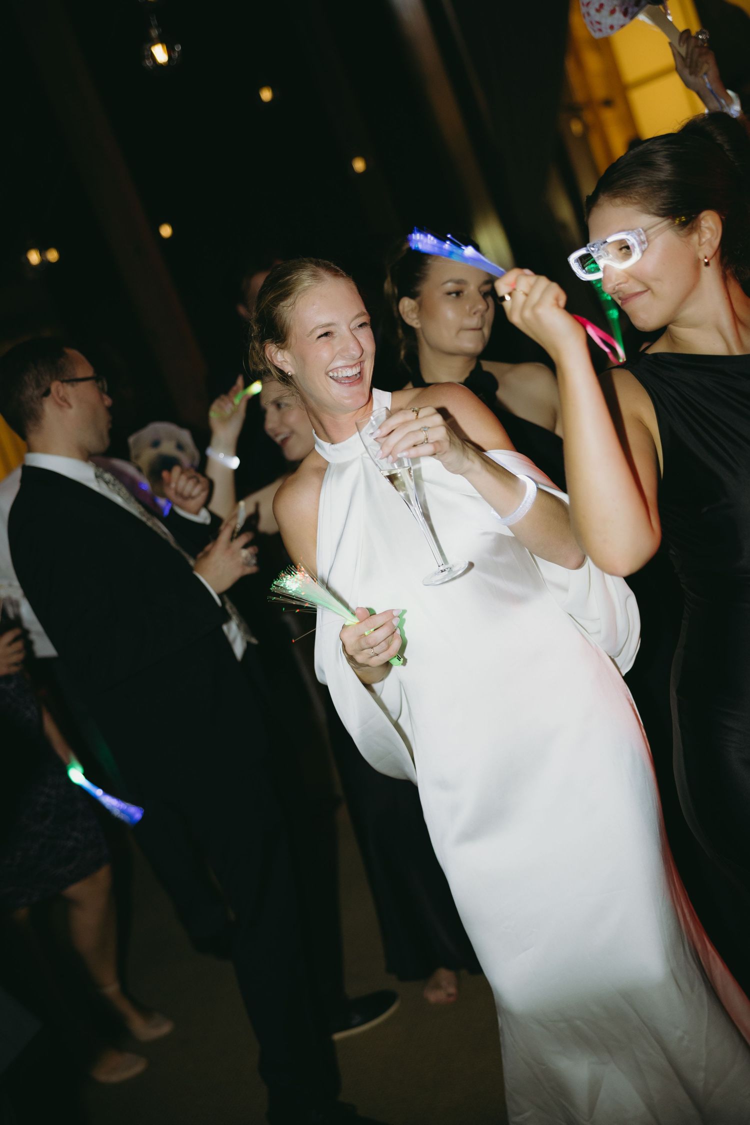 Laughing bride in white halter gown celebrates with guests holding glow sticks at lively wedding reception.