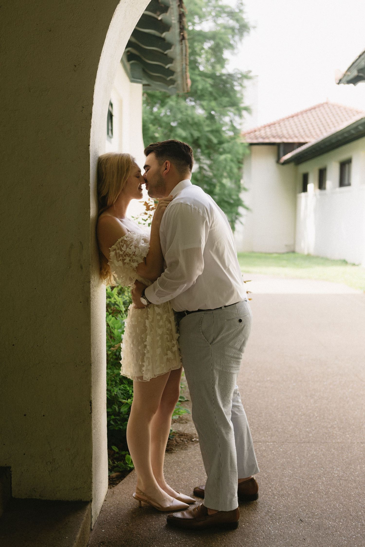 Couple shares romantic almost-kiss moment leaning against white archway column in elegant engagement photo session.