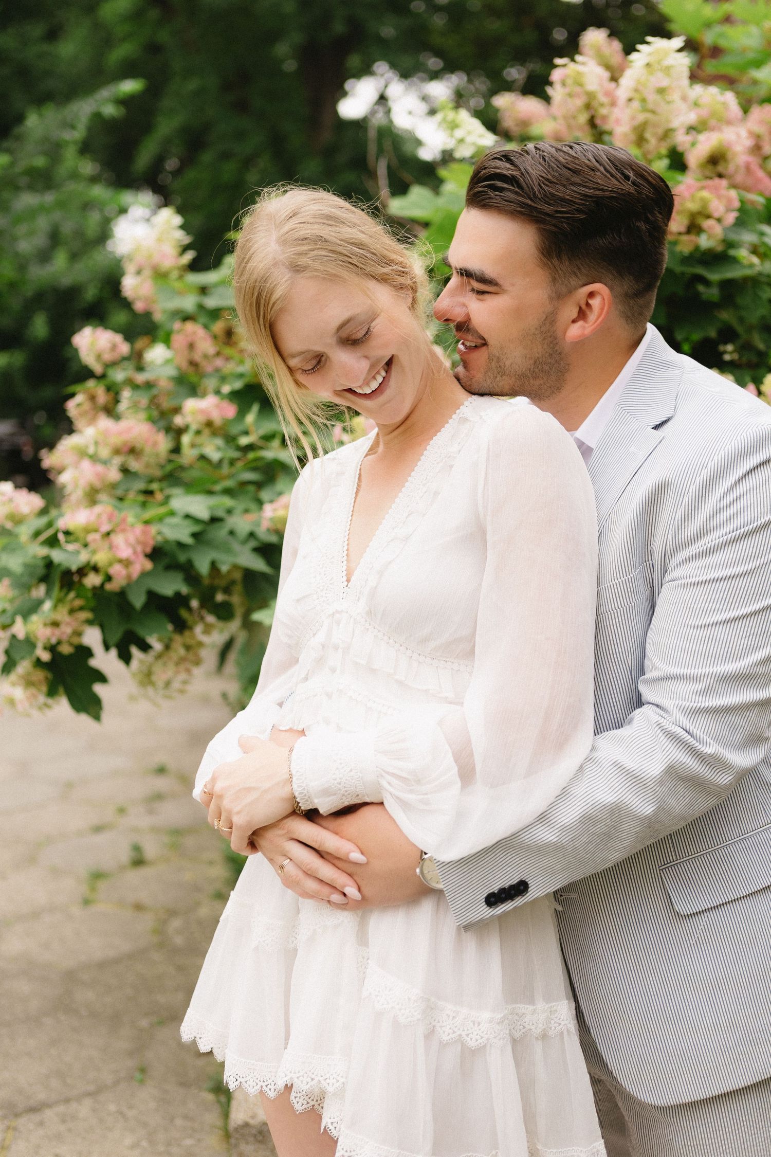 Smiling bride in white lace dress embraced by groom in gray suit among blooming hydrangeas.