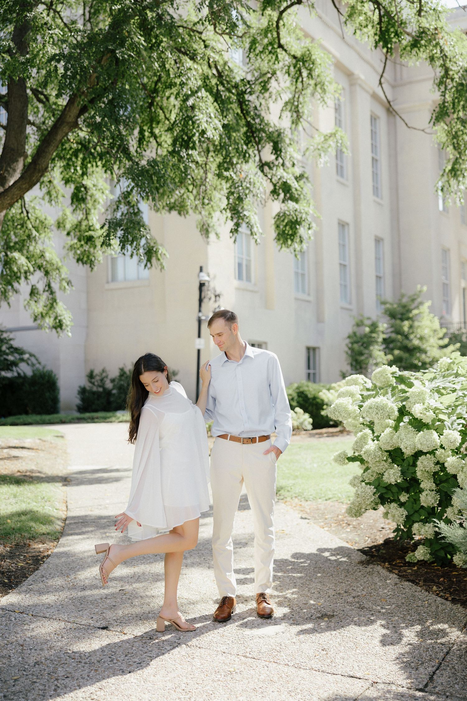 Couple walking together on a sunlit path beside white hydrangeas and a classic building exterior.