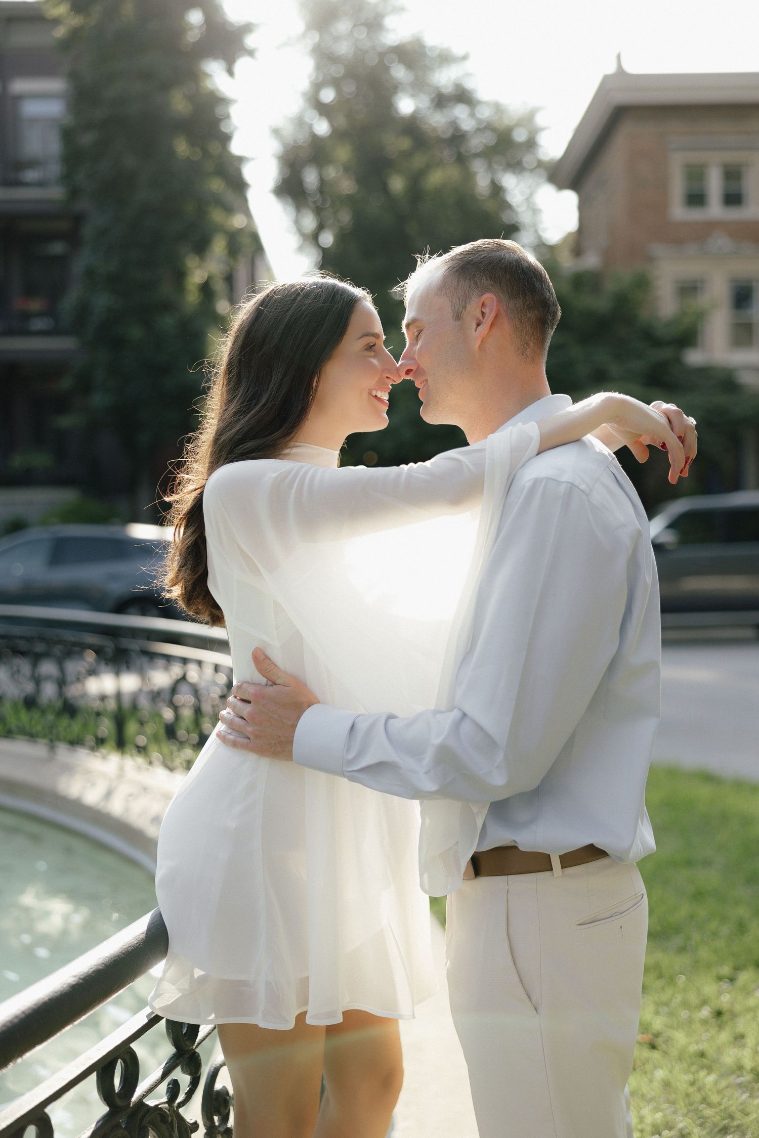 Smiling couple embracing near a fountain, woman in white dress with arms around man in light blue shirt.