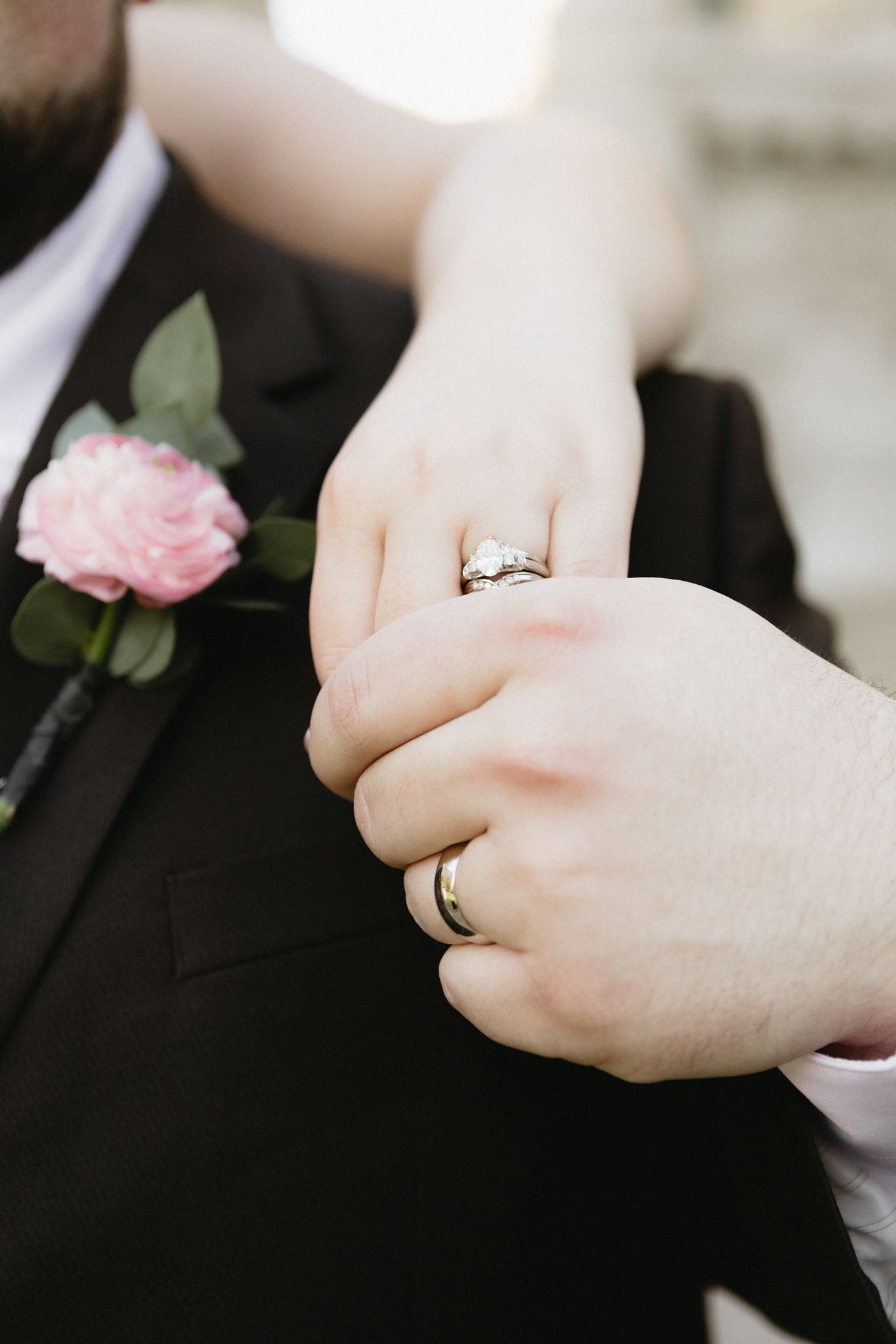 Close-up of wedding rings on hands with a pink rose boutonniere against a black suit.