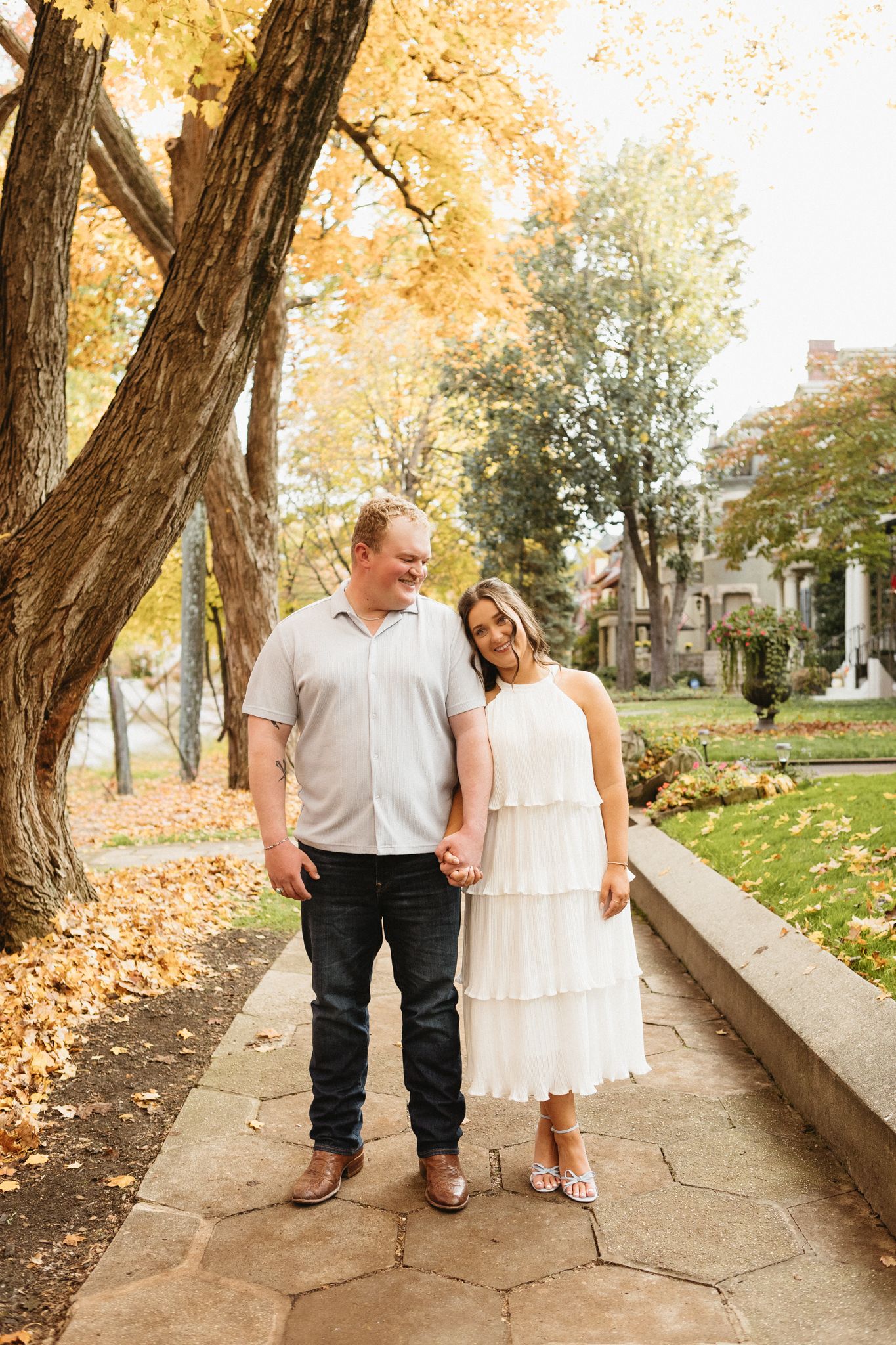A couple stands together on a stone path surrounded by autumn trees and historic architecture.
