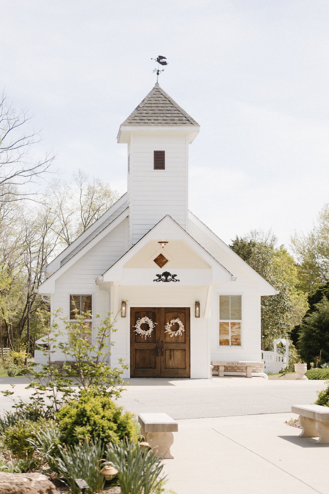 A charming white chapel with wooden double doors, decorative wreaths, and a tall steeple topped with a weathervane.