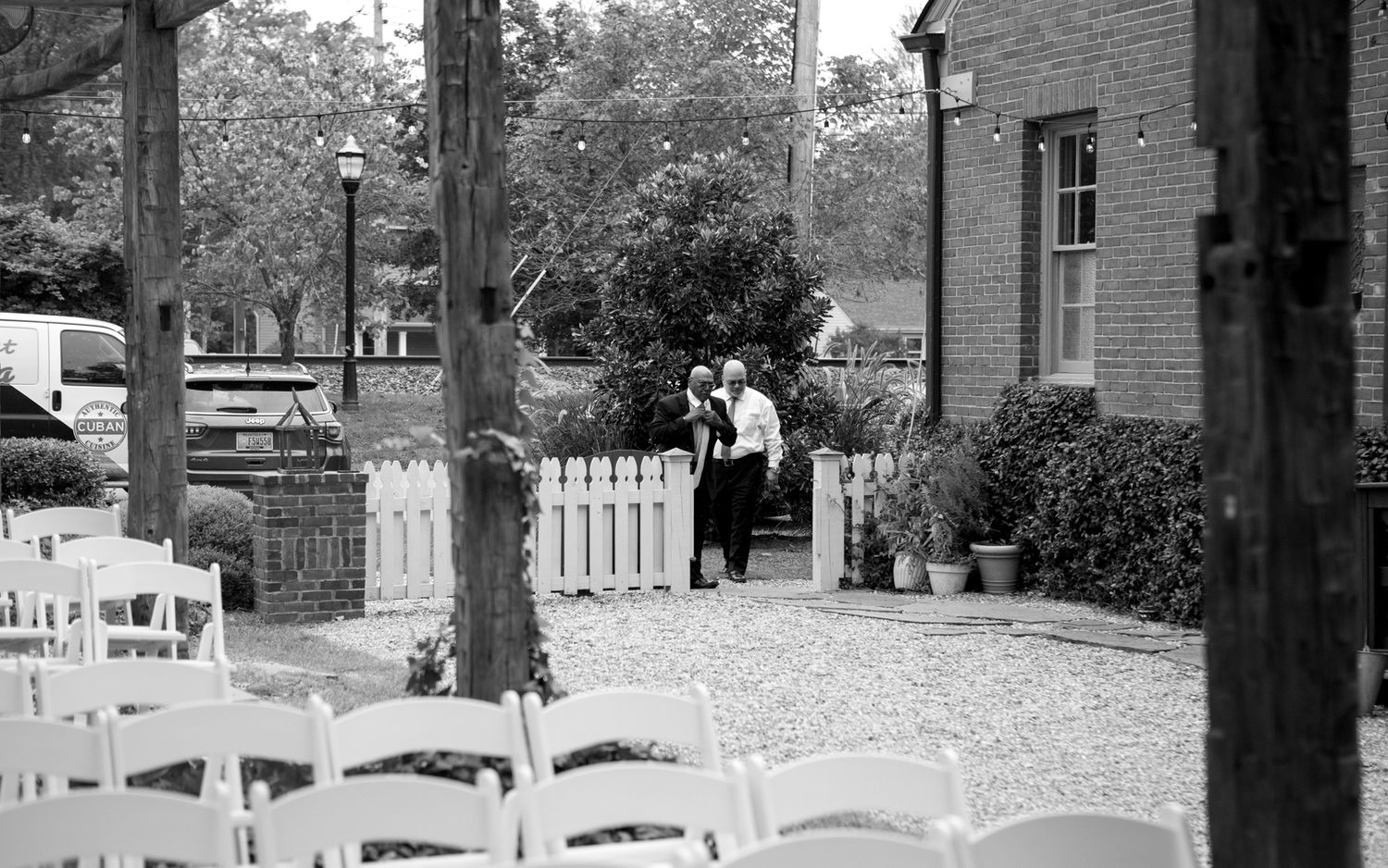 Black and white photograph showing a gravel path leading to a brick building with white chairs and garden setting.