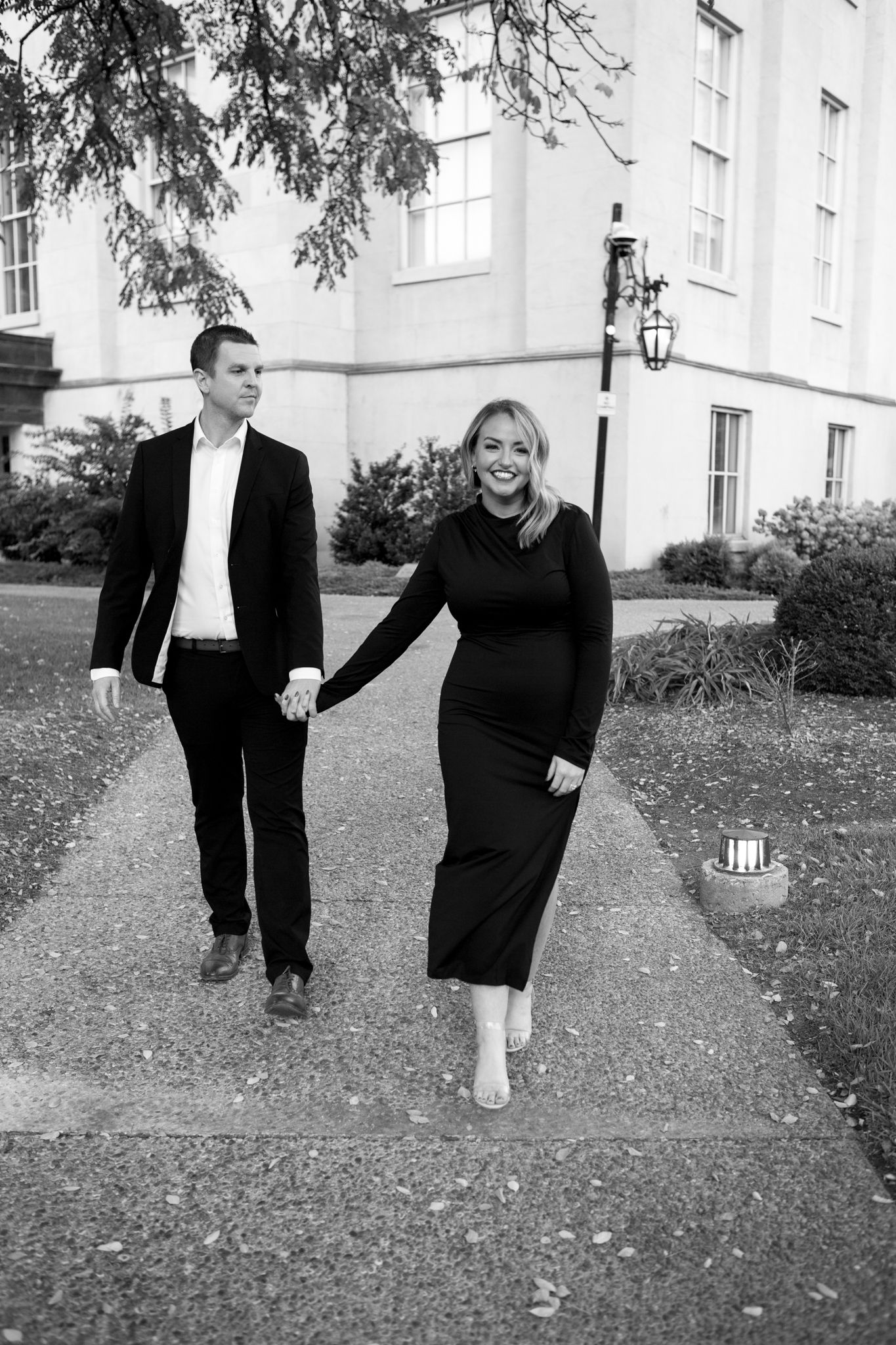 A black and white photograph of a couple walking hand in hand on a path near a elegant building.