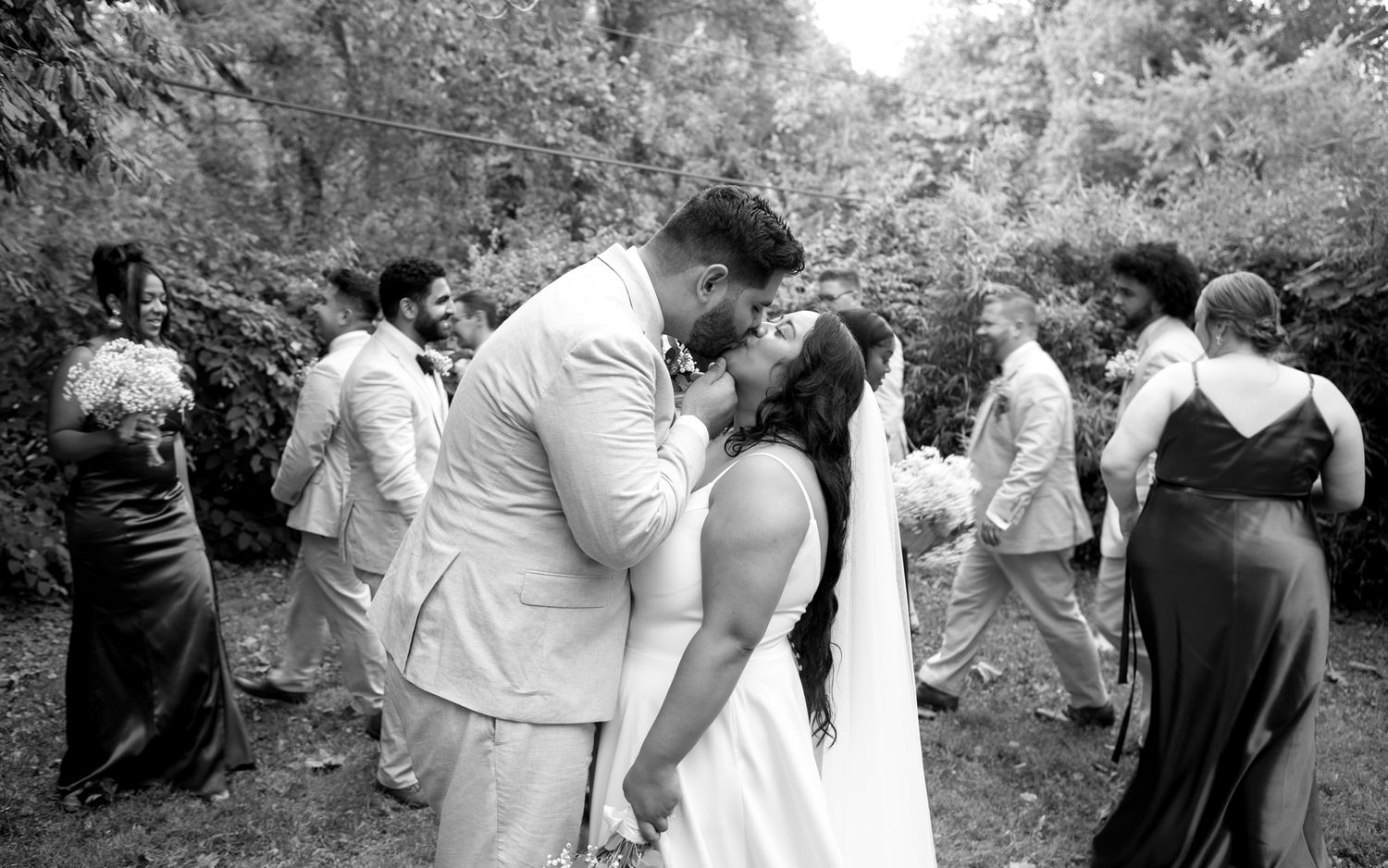 A couple shares an intimate kiss during an outdoor wedding ceremony while guests look on in black and white.
