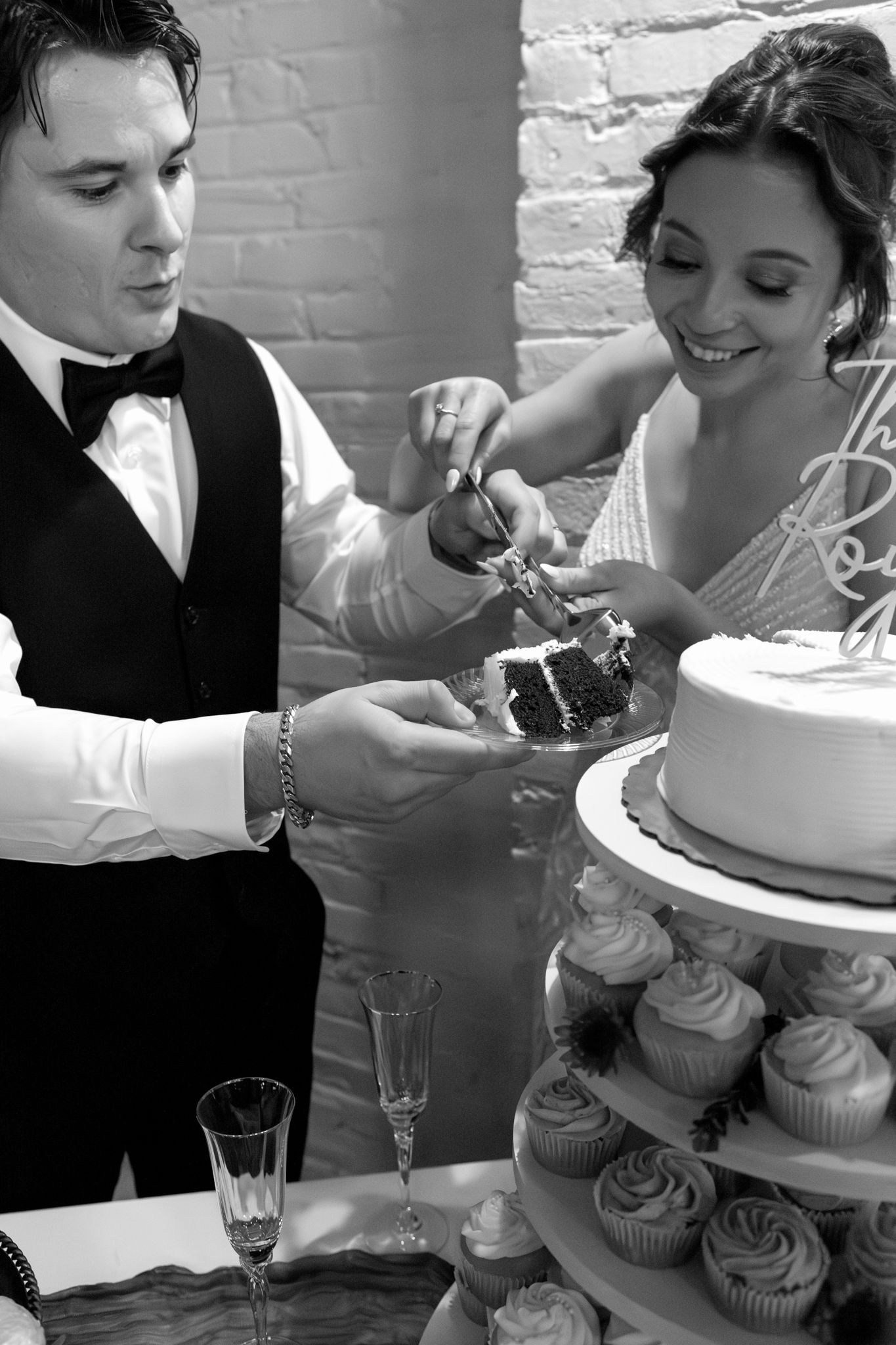A couple cuts their wedding cake together while standing next to a tiered cupcake display at their reception.