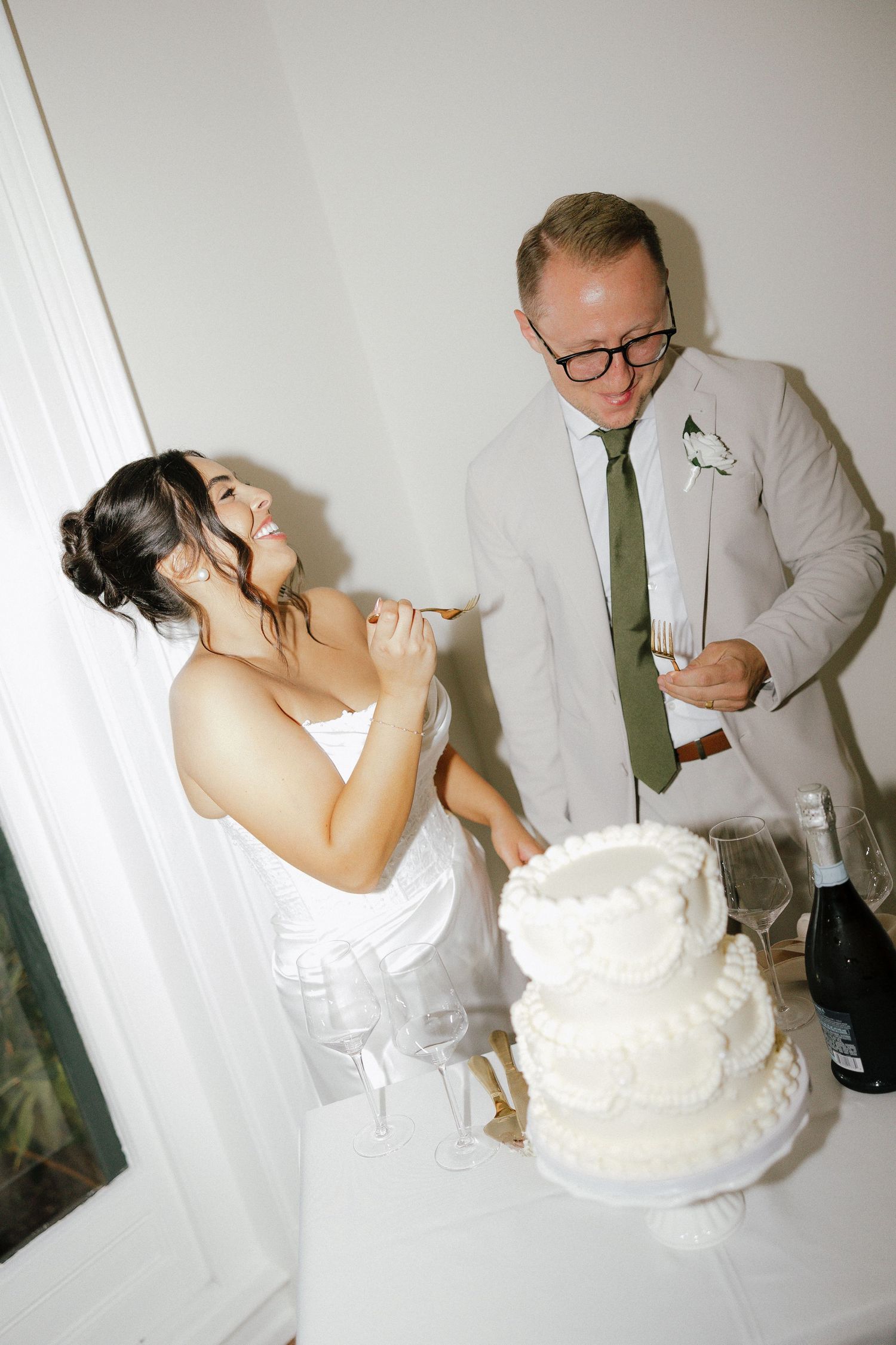 Laughing bride and groom cutting their two-tier white wedding cake together at their reception.