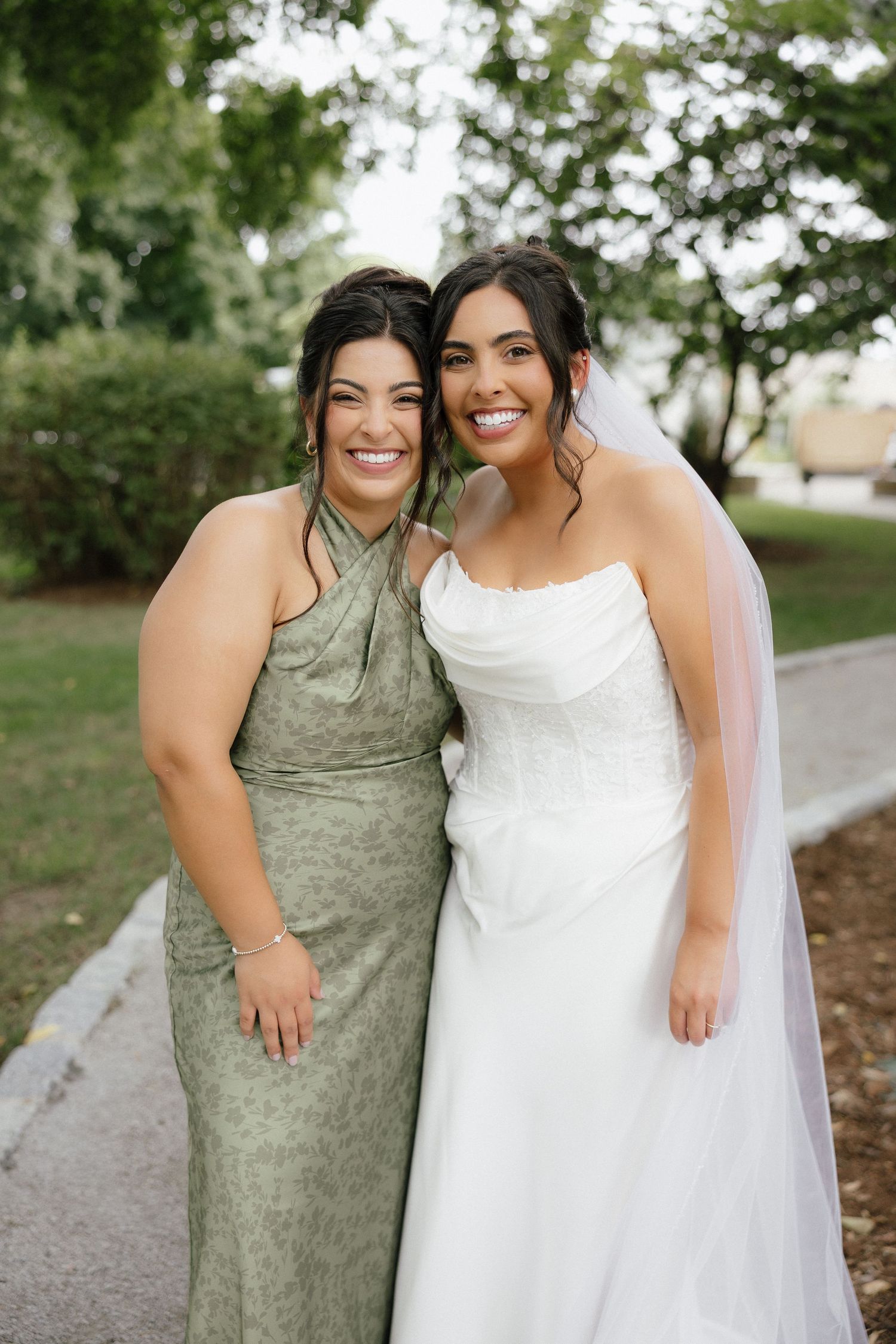 Bride in white strapless gown with veil smiles with bridesmaid in sage green dress outdoors.