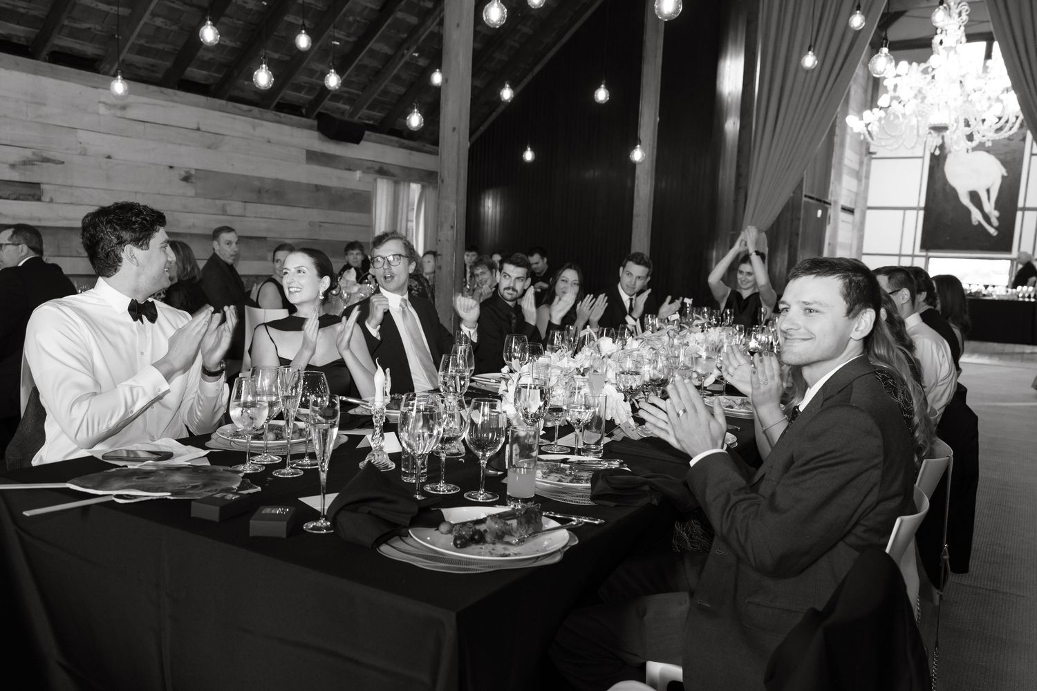 Black and white photo of wedding guests applauding at long reception table in rustic barn venue.