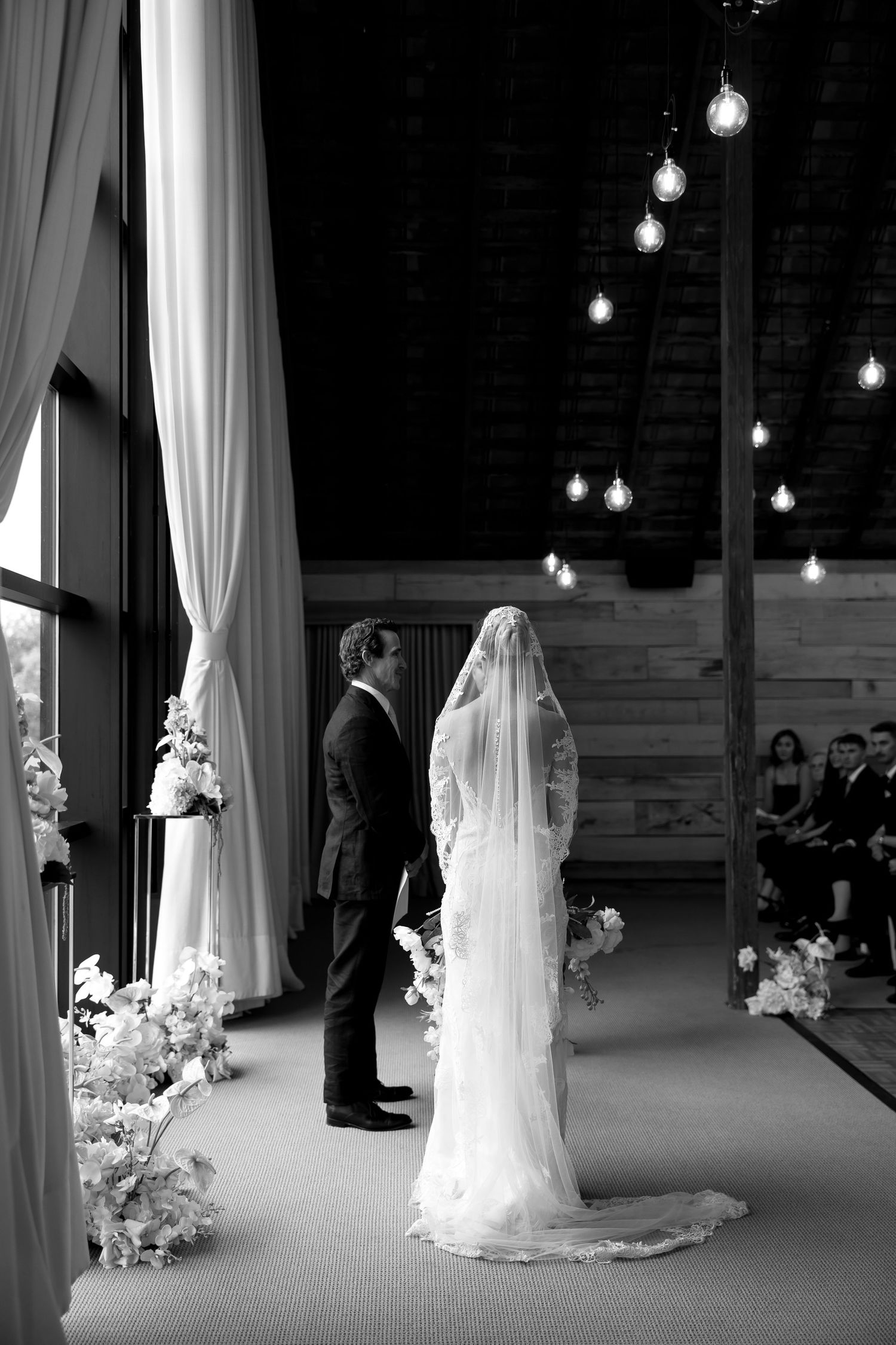 Bride and groom stand at the altar in a rustic barn wedding venue with hanging Edison bulb lights.