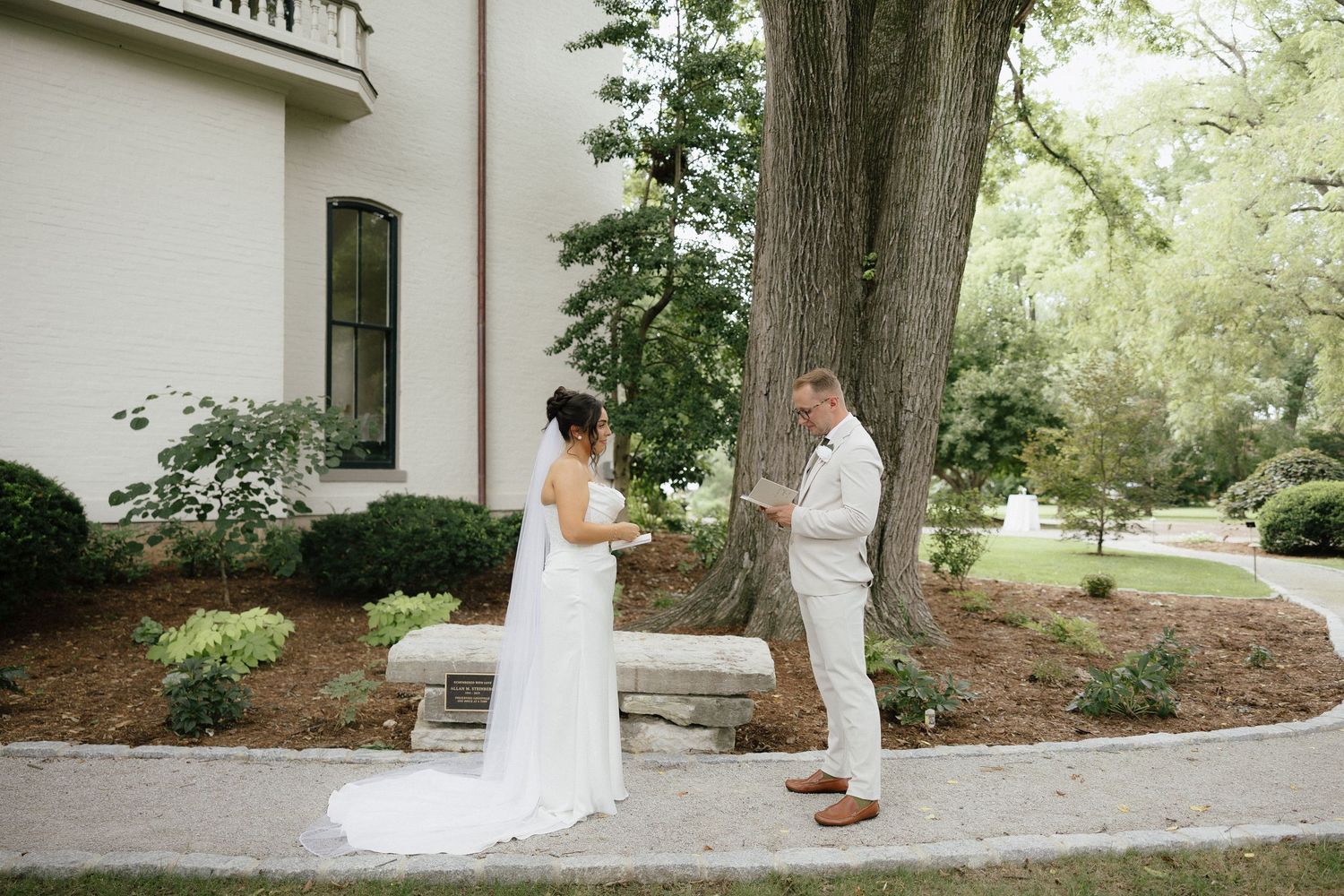Bride and groom share a first look moment beside a large tree outside a white wedding venue.
