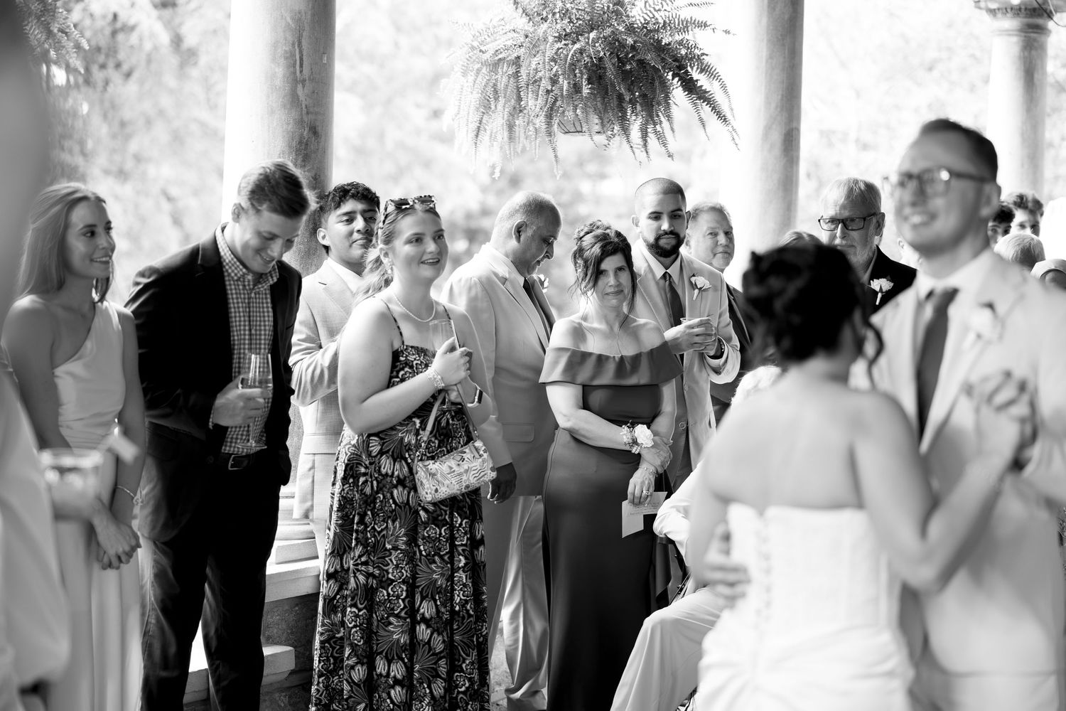 Wedding guests mingle and celebrate with drinks during outdoor reception under columned pavilion.