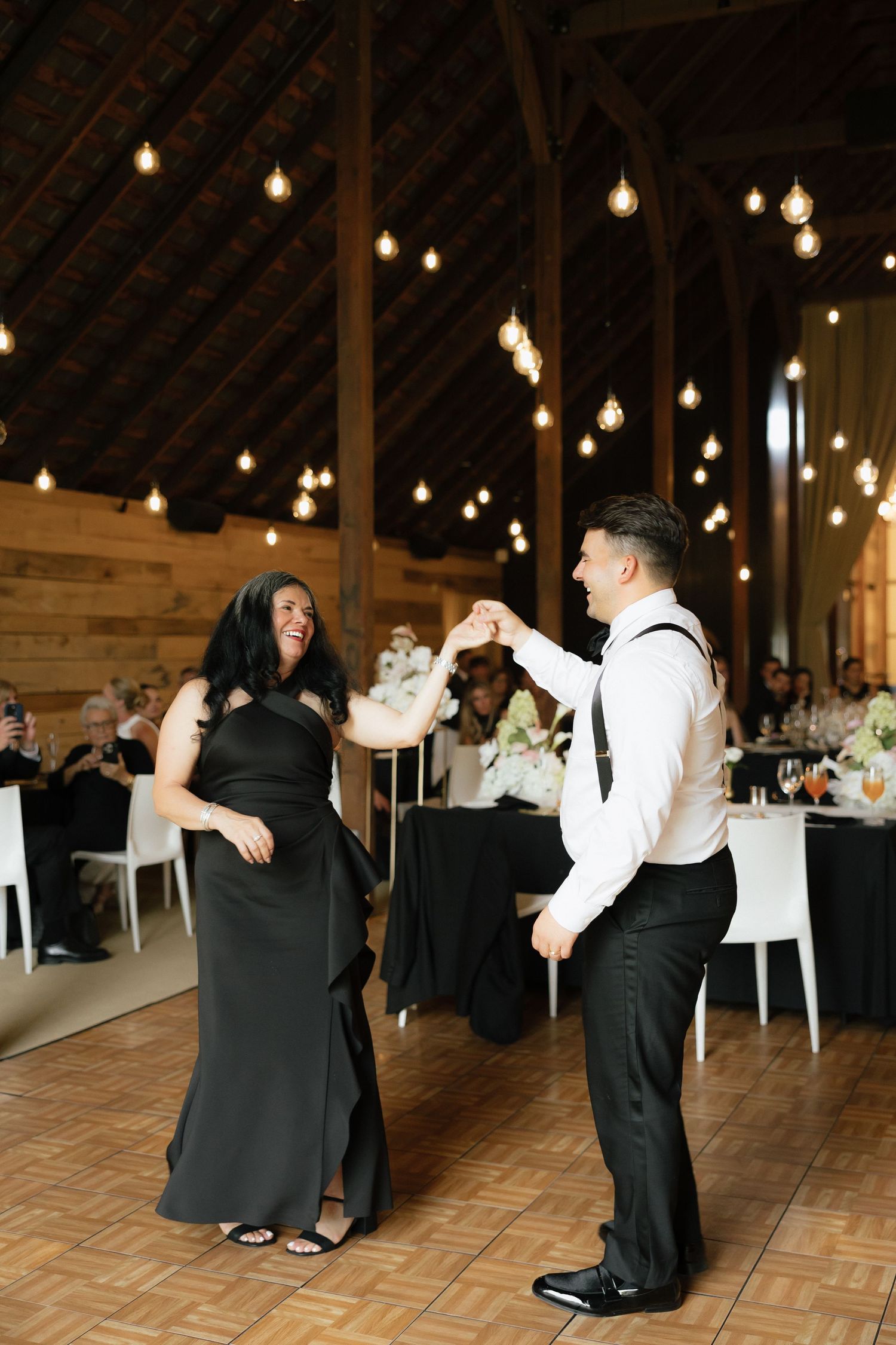 Groom dancing with mother in black dress on wooden dance floor at barn wedding reception.