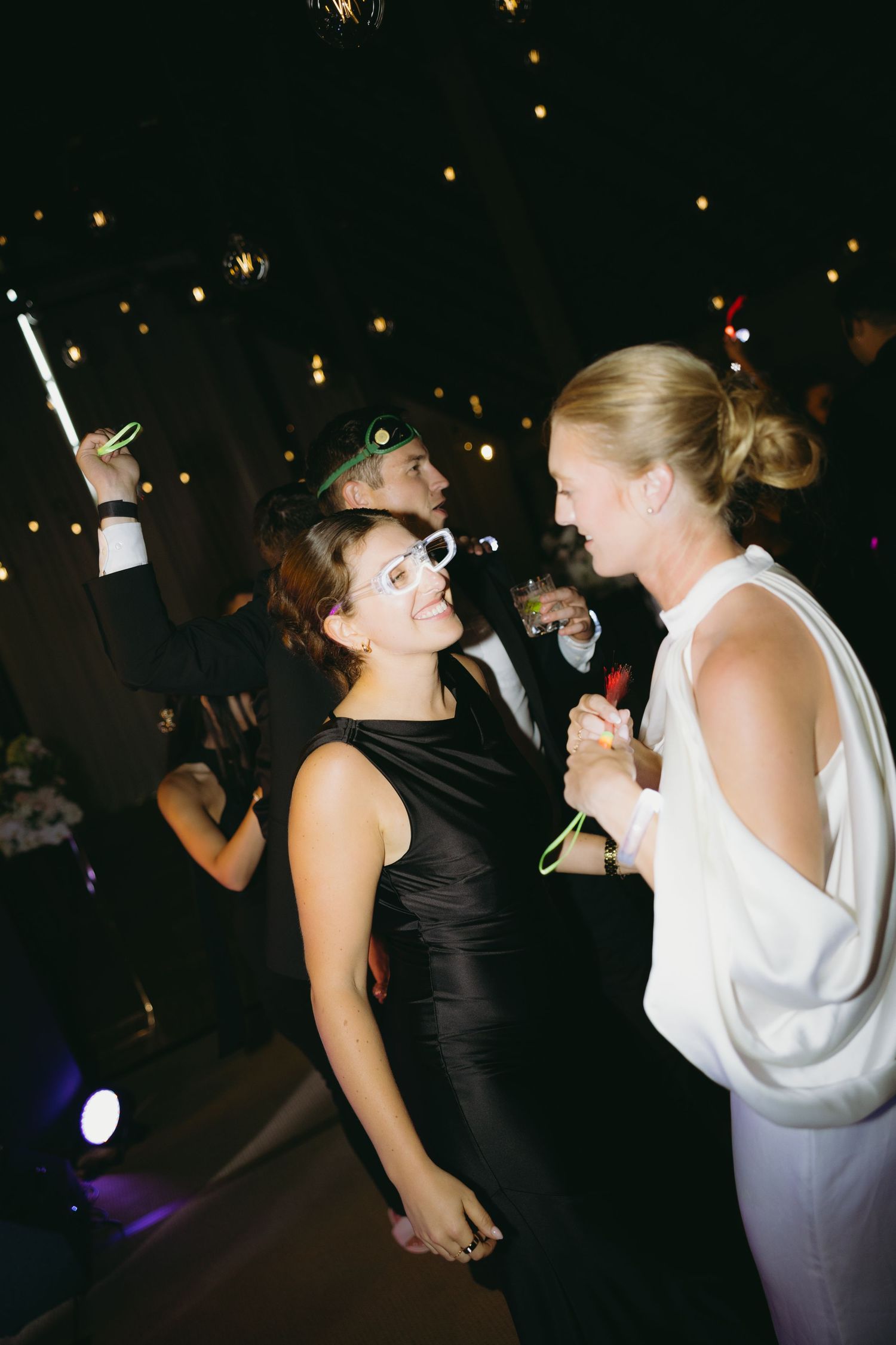 Bride in white gown and guest in black dress dance together with glow sticks at evening reception.