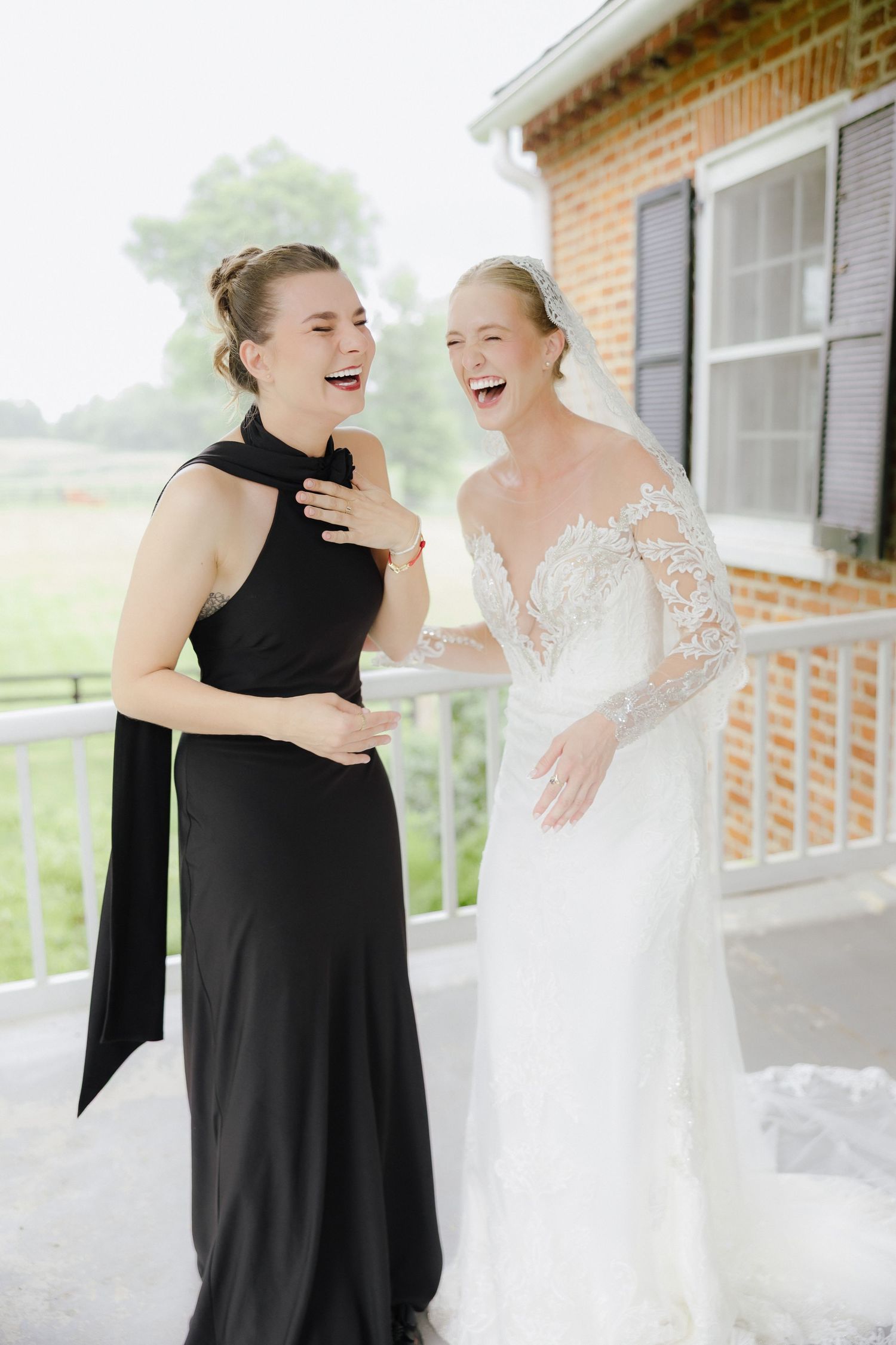 Bride in lace gown laughing joyfully with bridesmaid in black dress on a porch.
