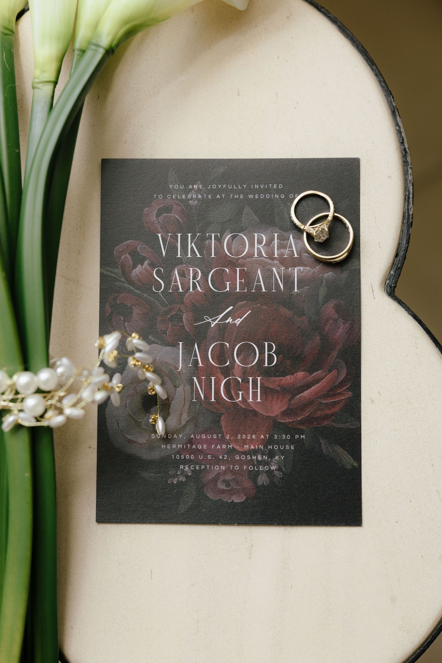 Dark floral wedding invitation for Viktoria Sargeant and Jacob Nigh with gold rings resting on top.