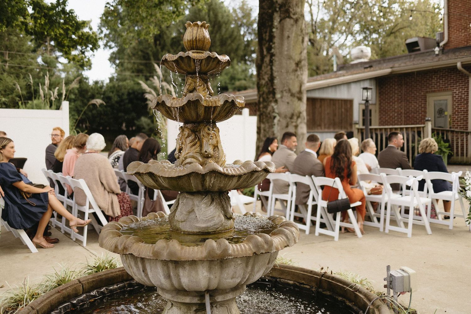 Outdoor wedding ceremony setup with white chairs and stone fountain in garden setting.