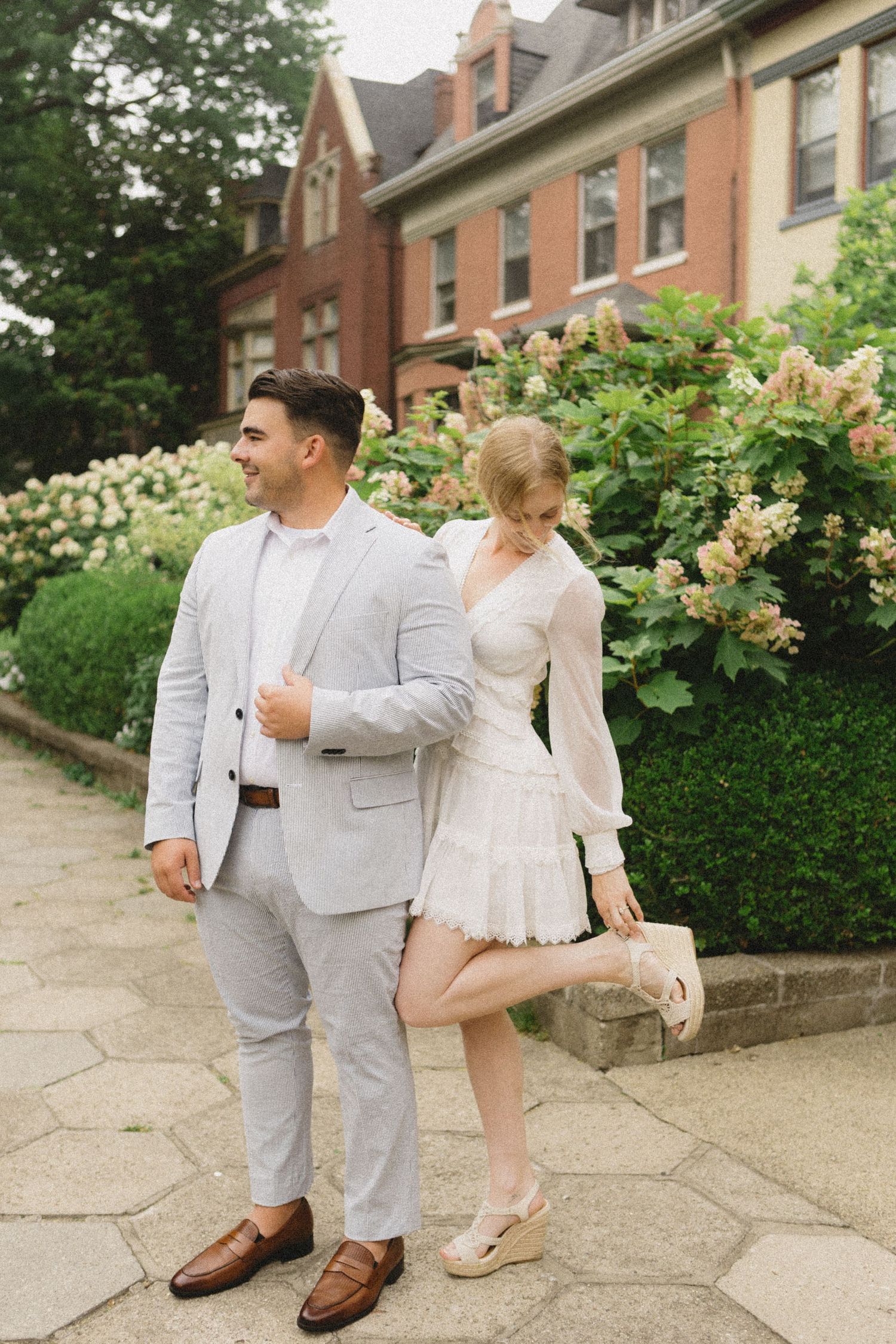 Engaged couple posing outdoors in white outfits before a brick building with hydrangea bushes.
