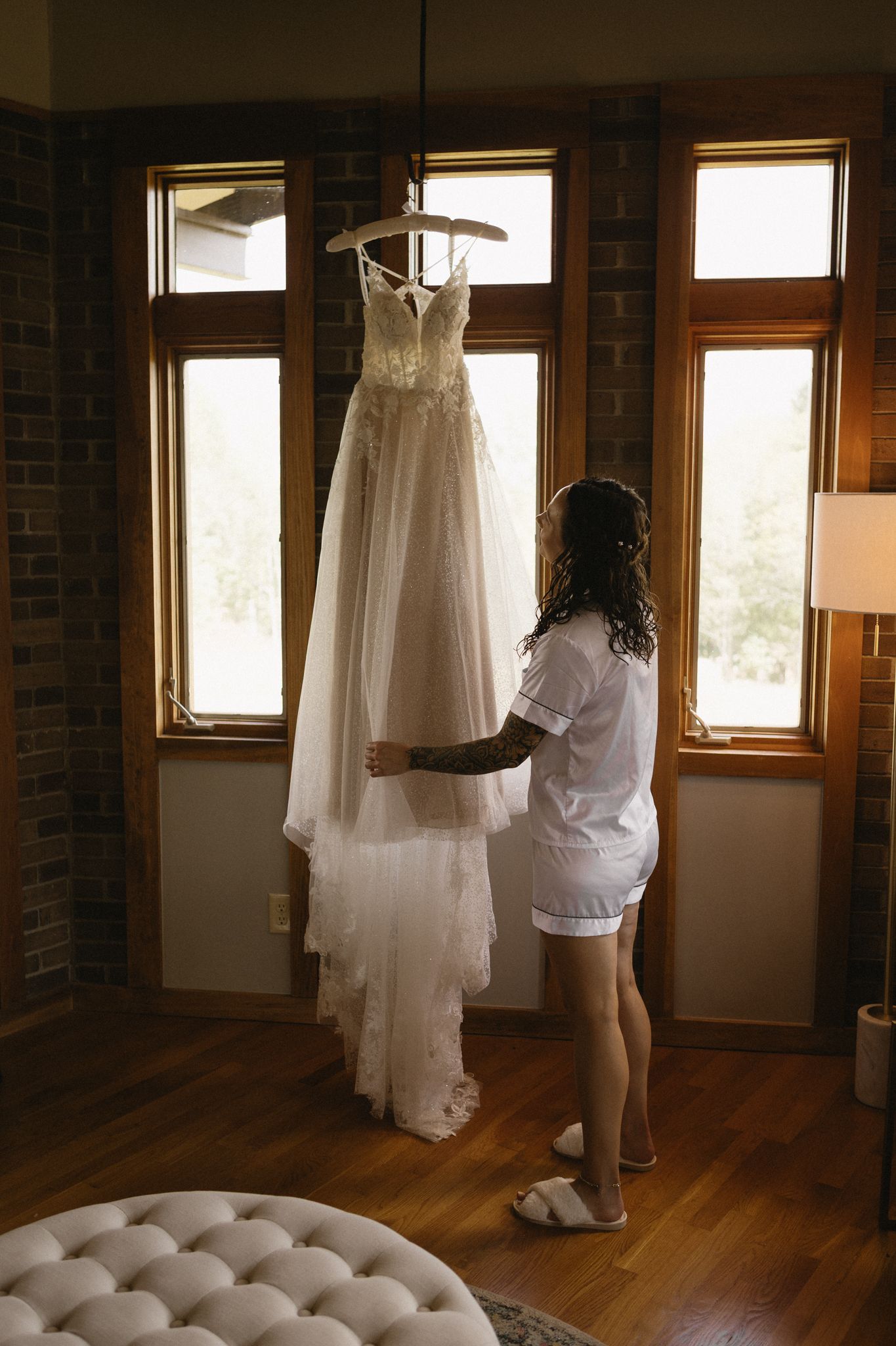 Wedding dress and white robe hanging by sunlit windows in rustic room with hardwood floors and exposed brick walls.